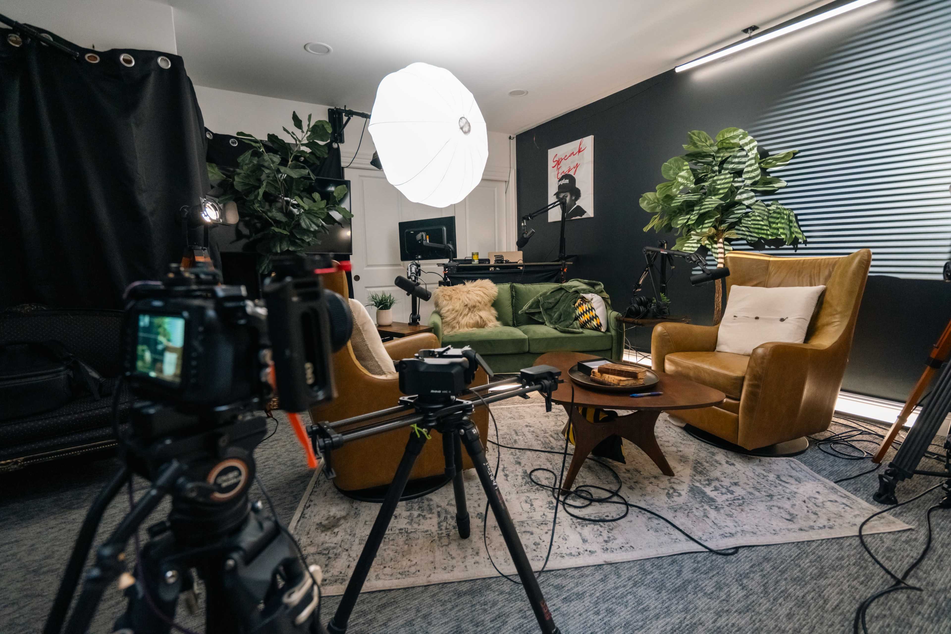 A well-arranged indoor studio setup with a green sofa, two armchairs, and various recording equipment, surrounded by plants and posters.