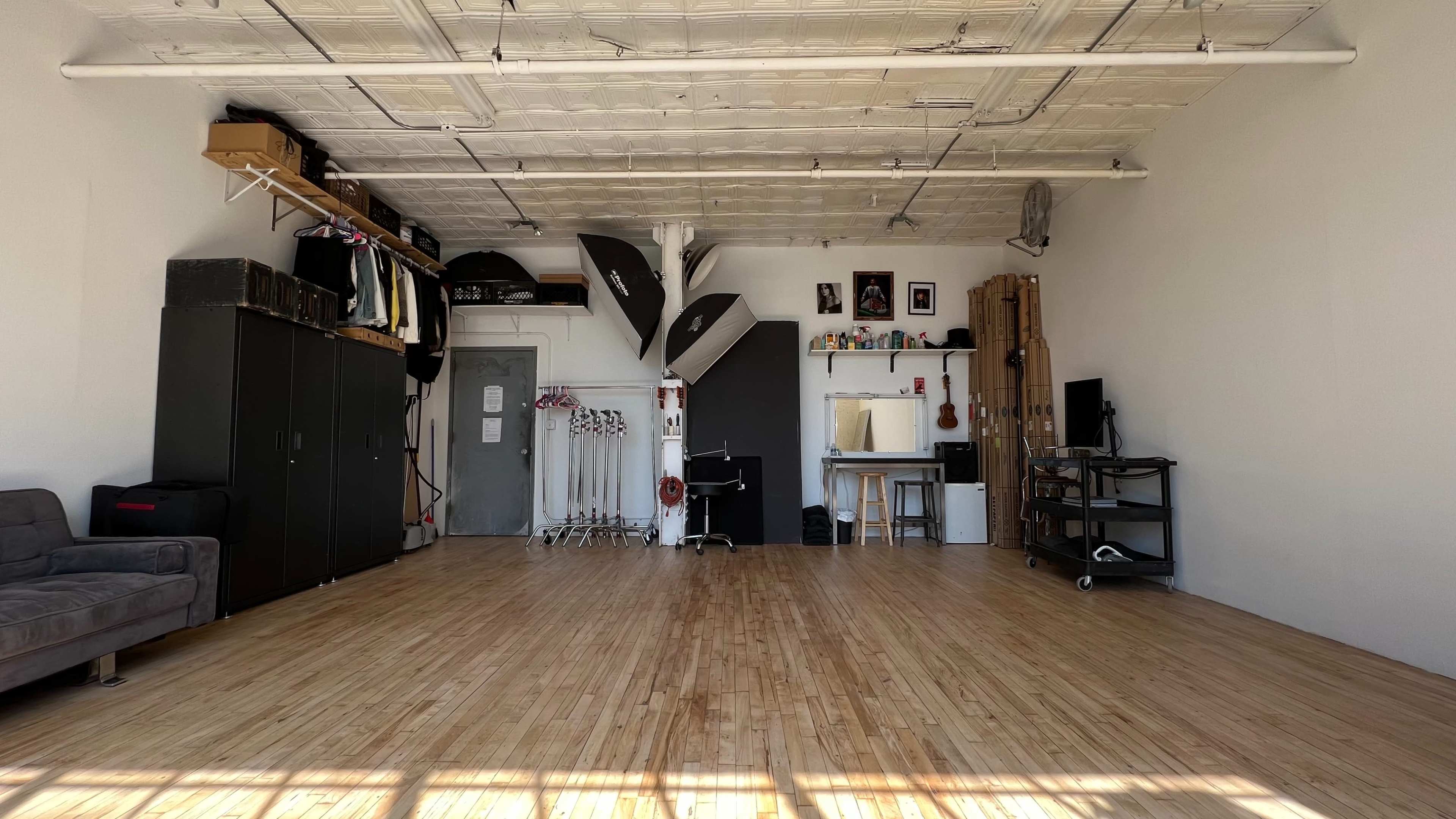 The image shows a spacious, empty studio with wooden flooring, storage cabinets, photography equipment, and furniture along the walls.
