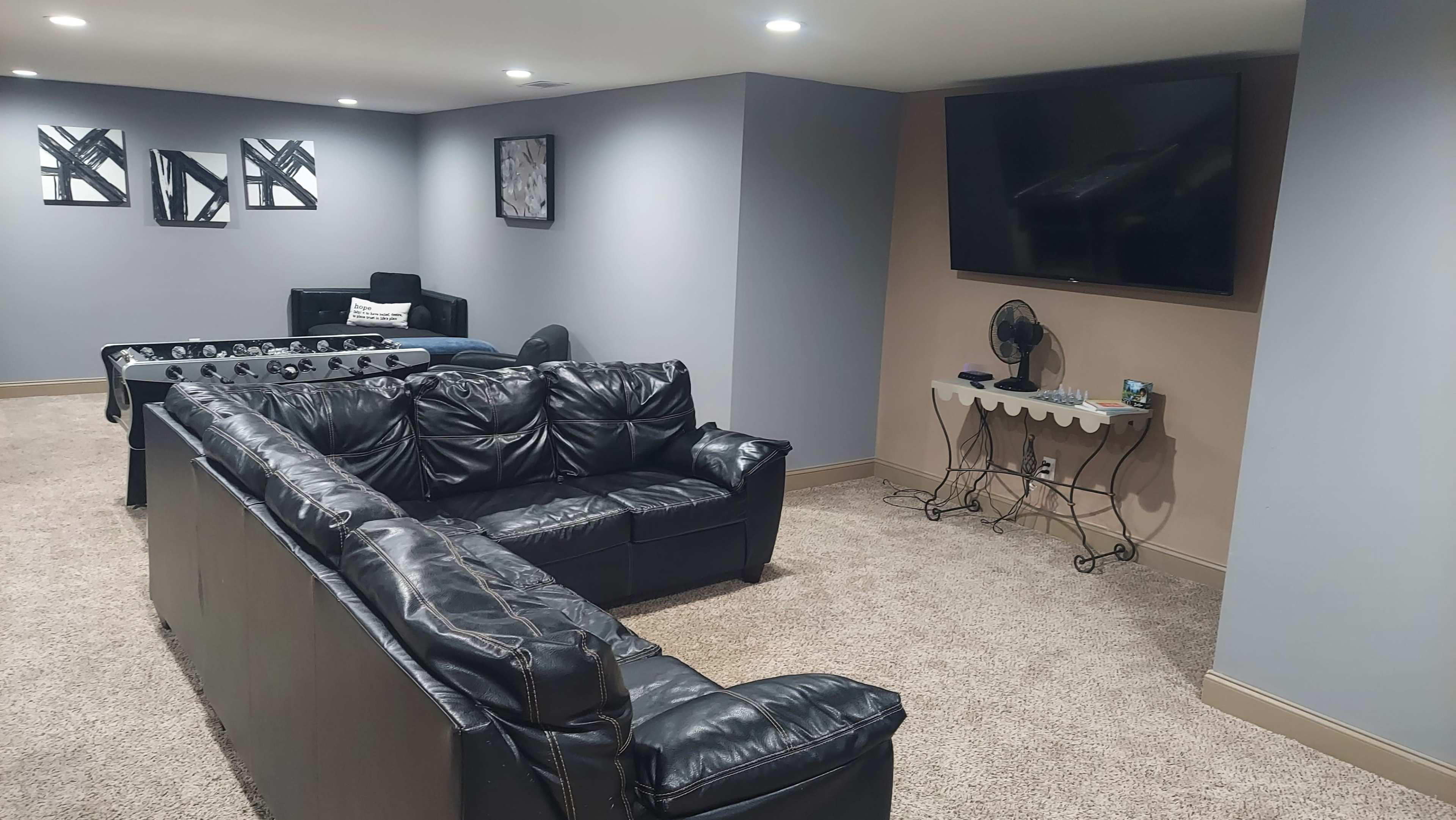 A spacious basement room features a black leather sectional couch, a foosball table, and a wall-mounted television.