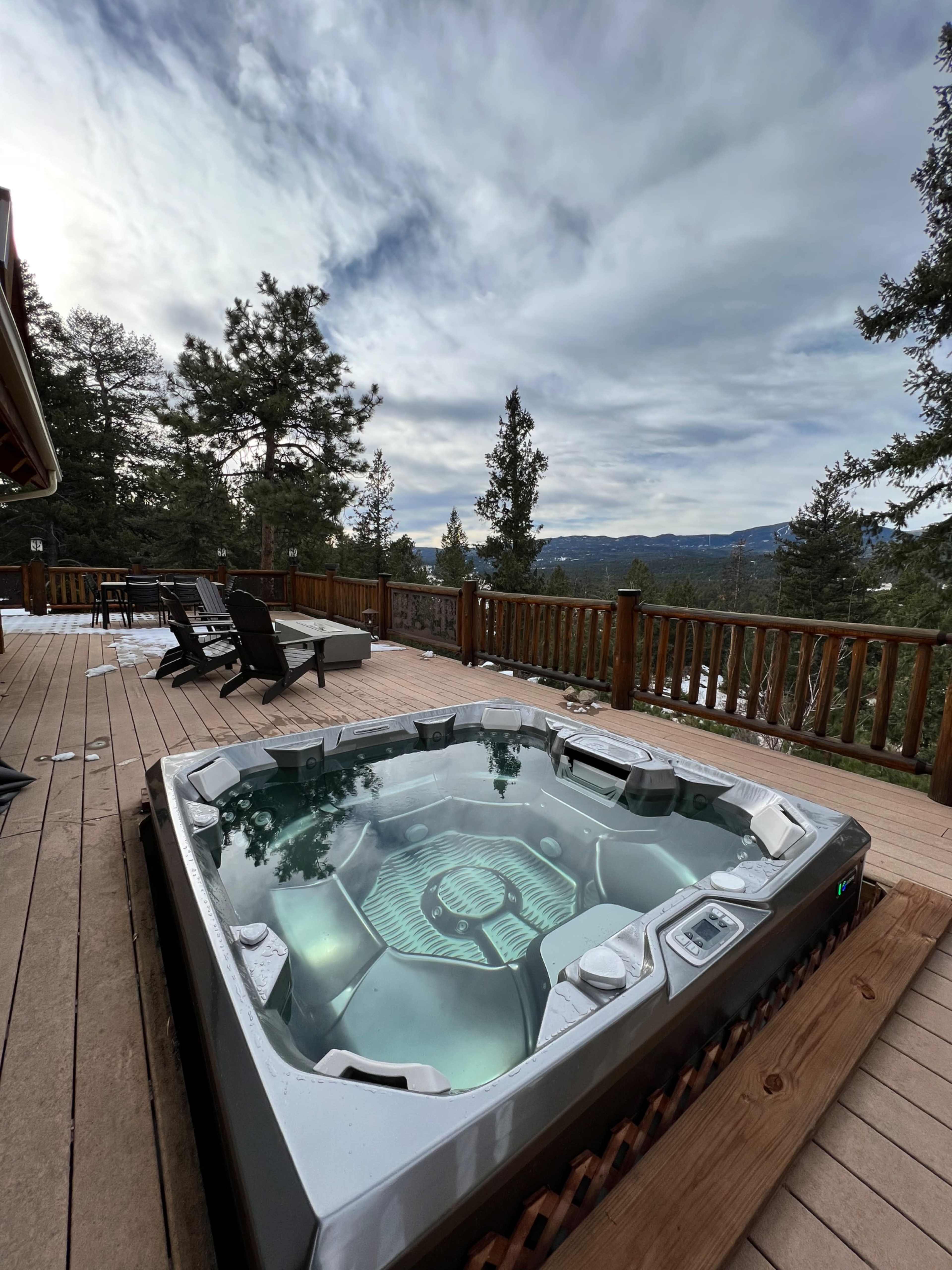 A hot tub is situated on a wooden deck overlooking a forested landscape and distant hills under a cloudy sky.
