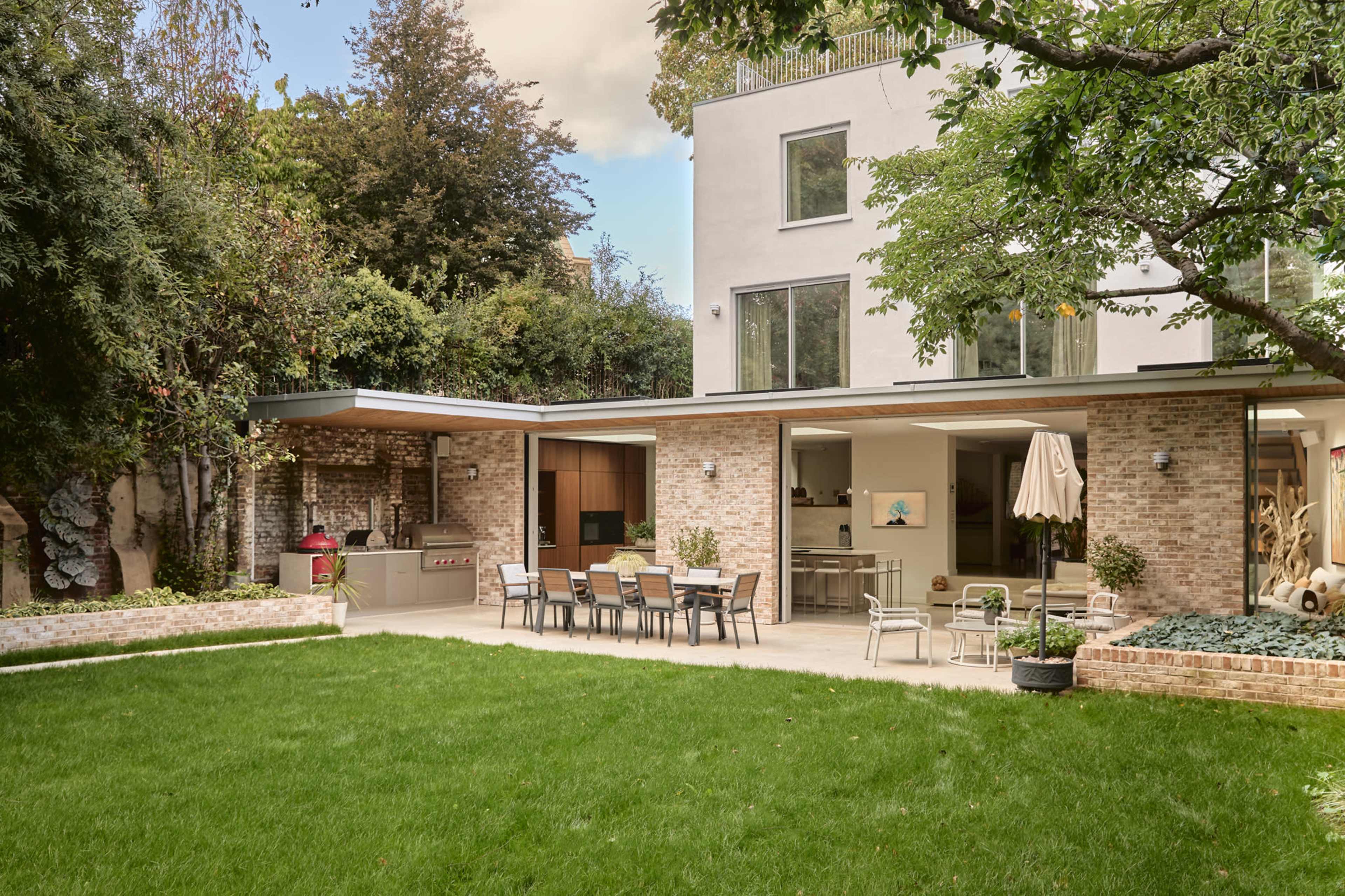 A modern residential backyard features a well-manicured lawn, a spacious patio area with dining furniture, and an outdoor kitchen adjacent to a contemporary house with large windows.