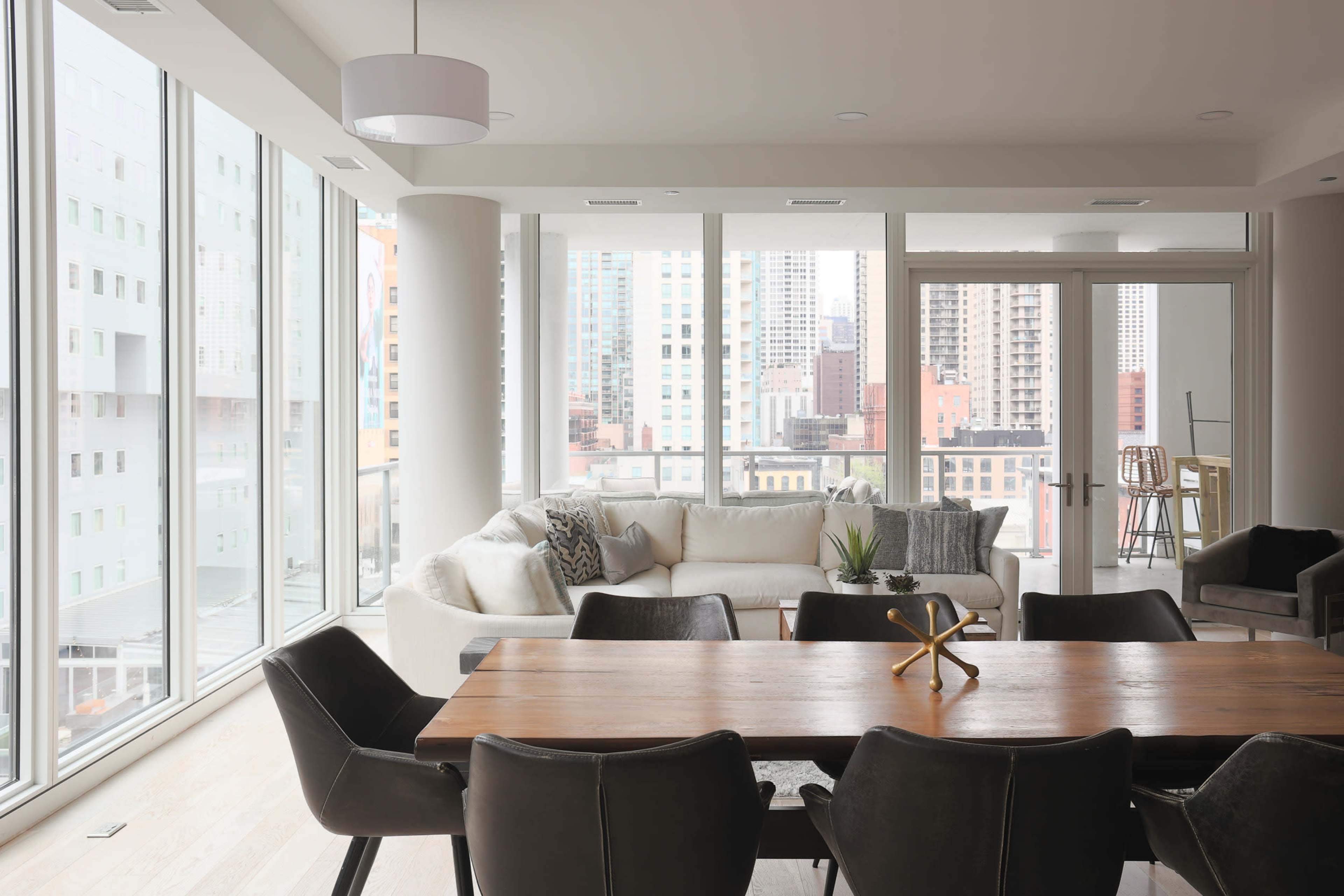 A modern living space features a large dining table, a light-colored sofa, and expansive windows showcasing a city skyline.