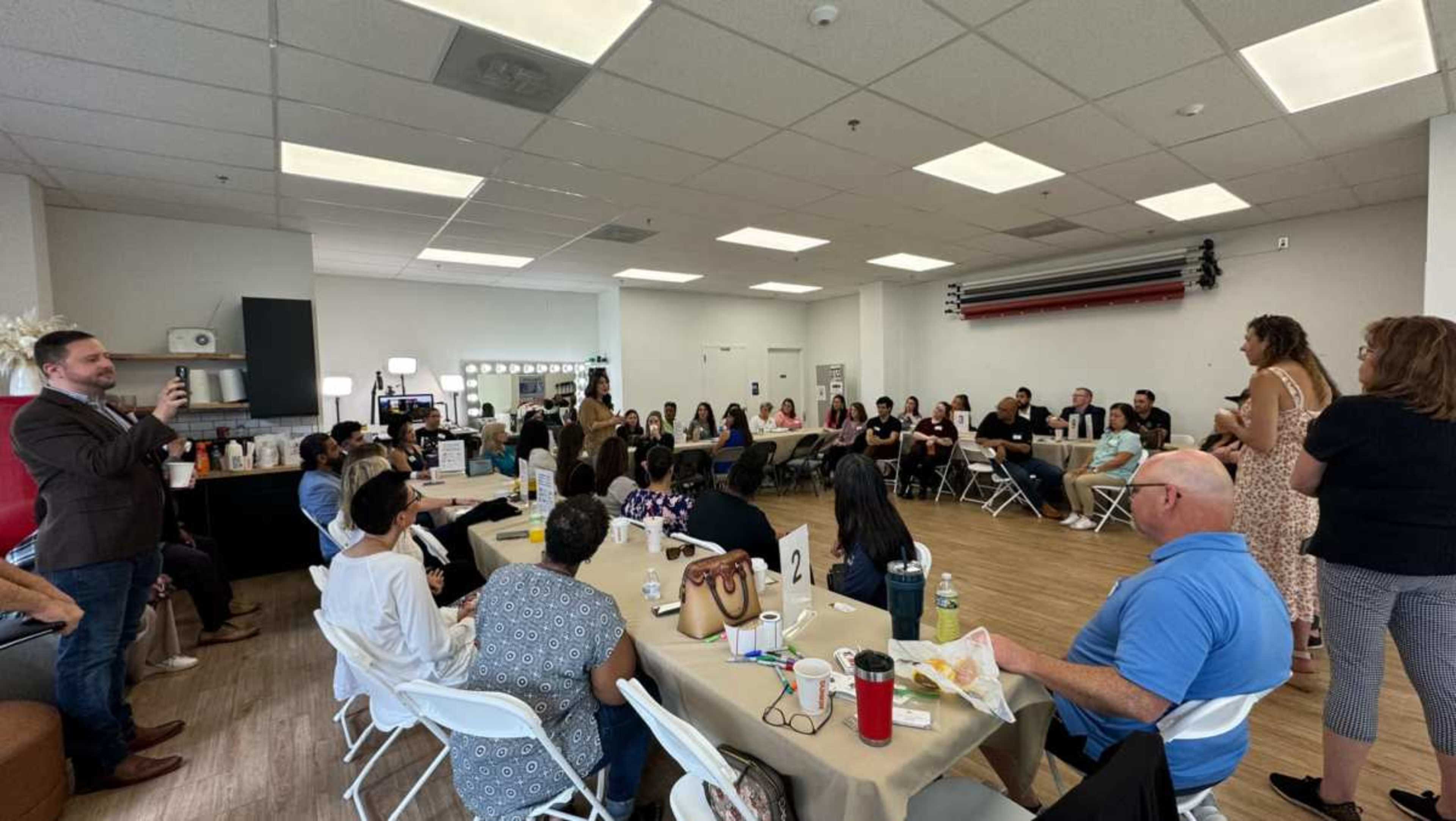 A large group of people is gathered in a well-lit room, sitting at tables and listening to a speaker at the front.