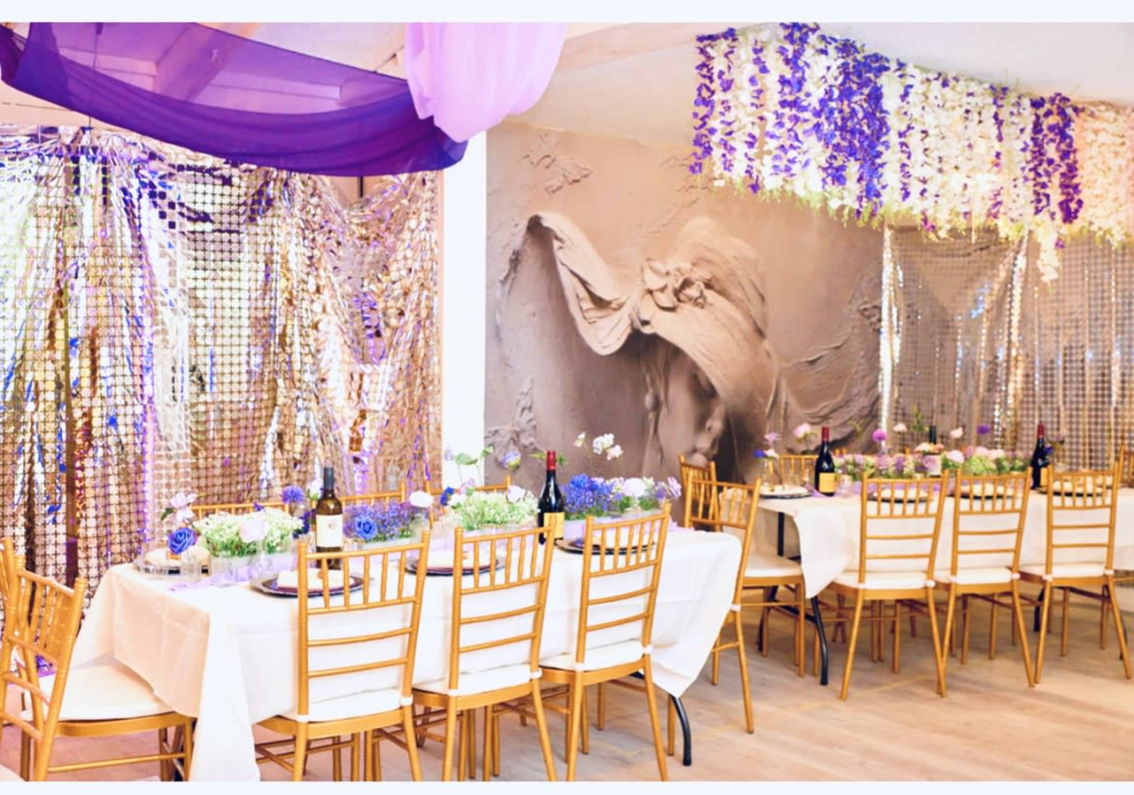 The image shows a decorated dining area with a long table set with white tablecloths and gold chairs, adorned with floral centerpieces, sparkling backdrops, and draped fabric.