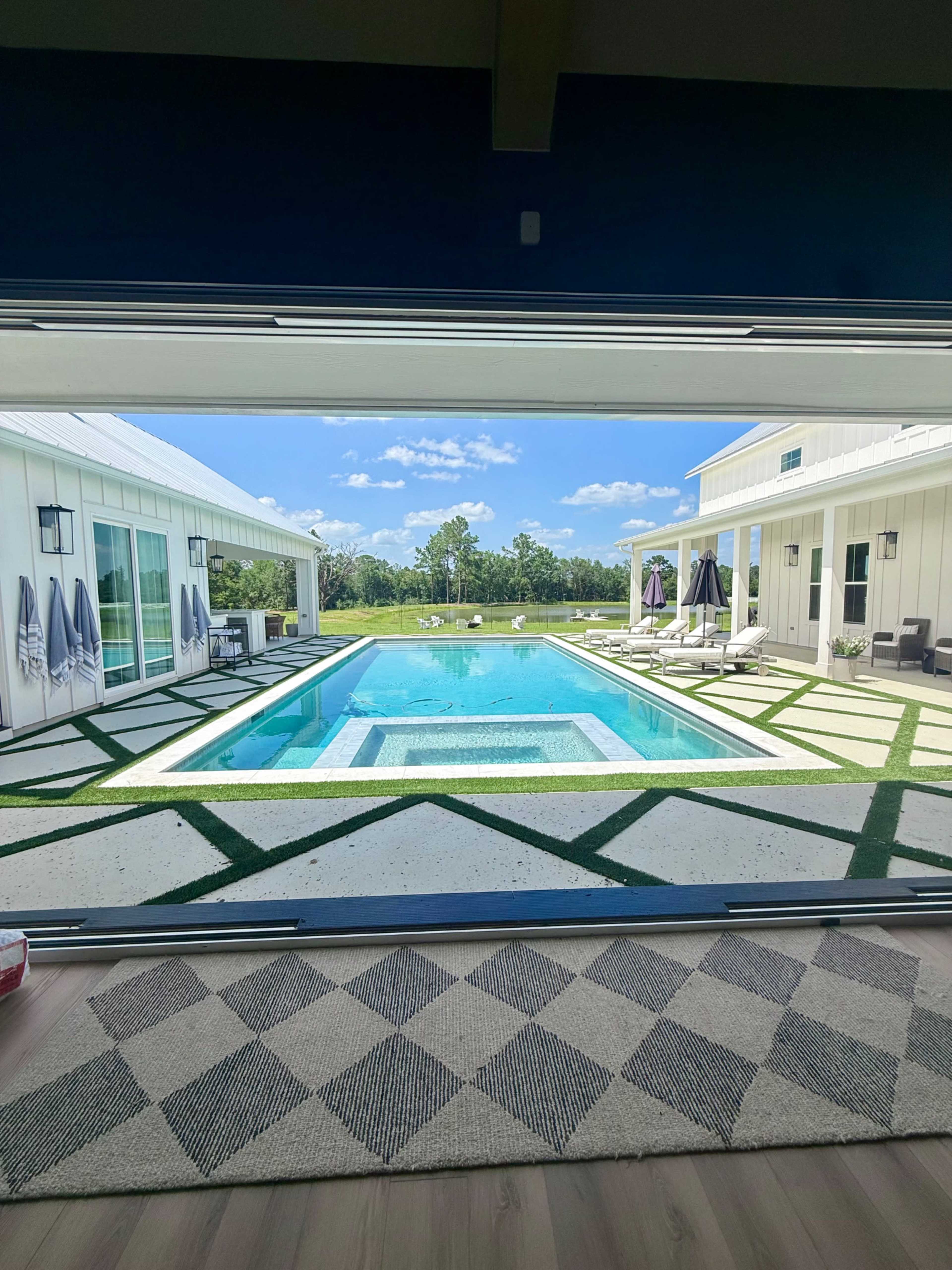 The image shows a view of a modern home with a large swimming pool surrounded by green landscaping and patio furniture.