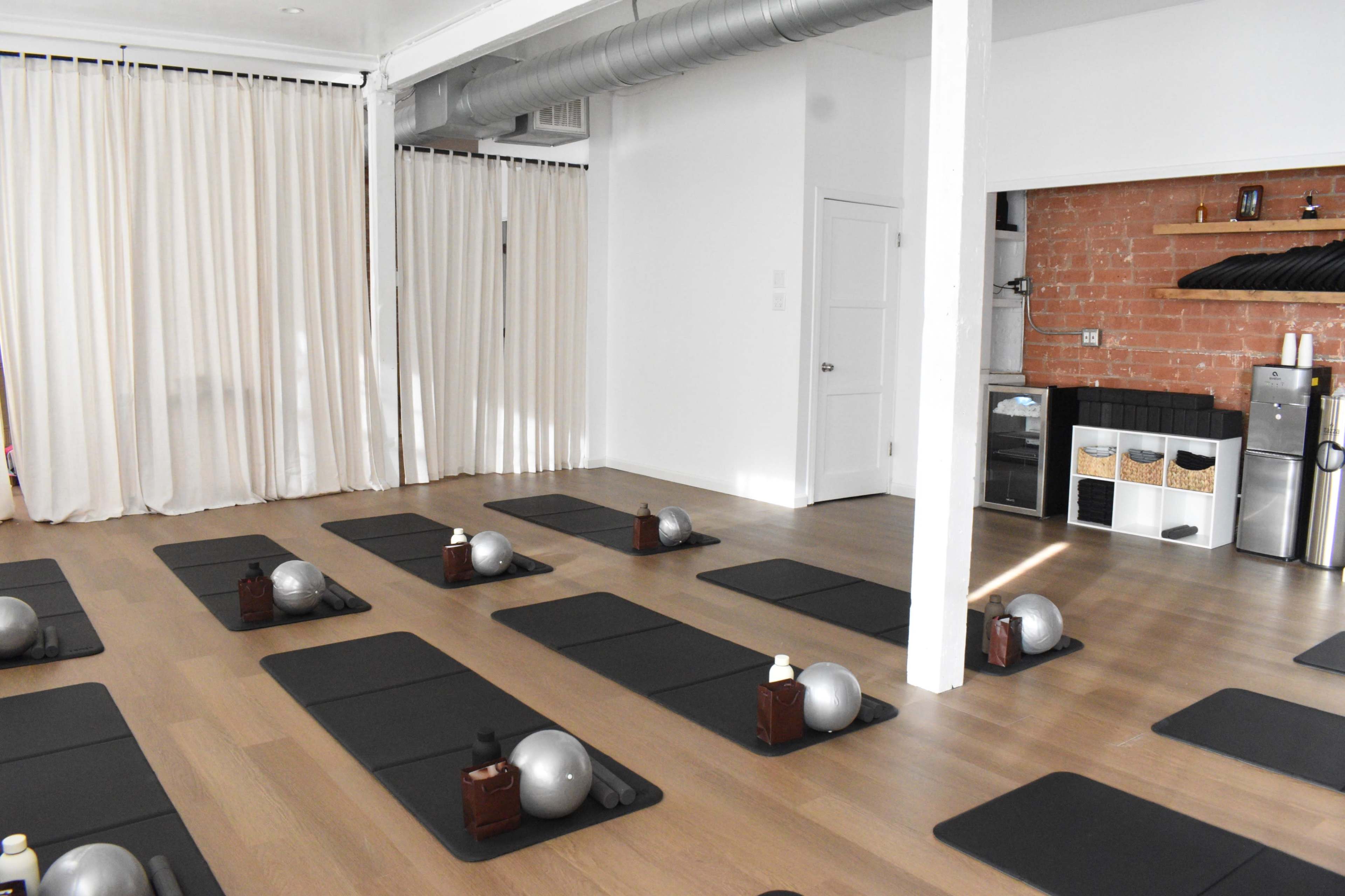 The image shows a minimalist exercise studio with several yoga mats arranged in rows, each featuring a small ball and a bottle on top, along with a kitchenette area in the background.