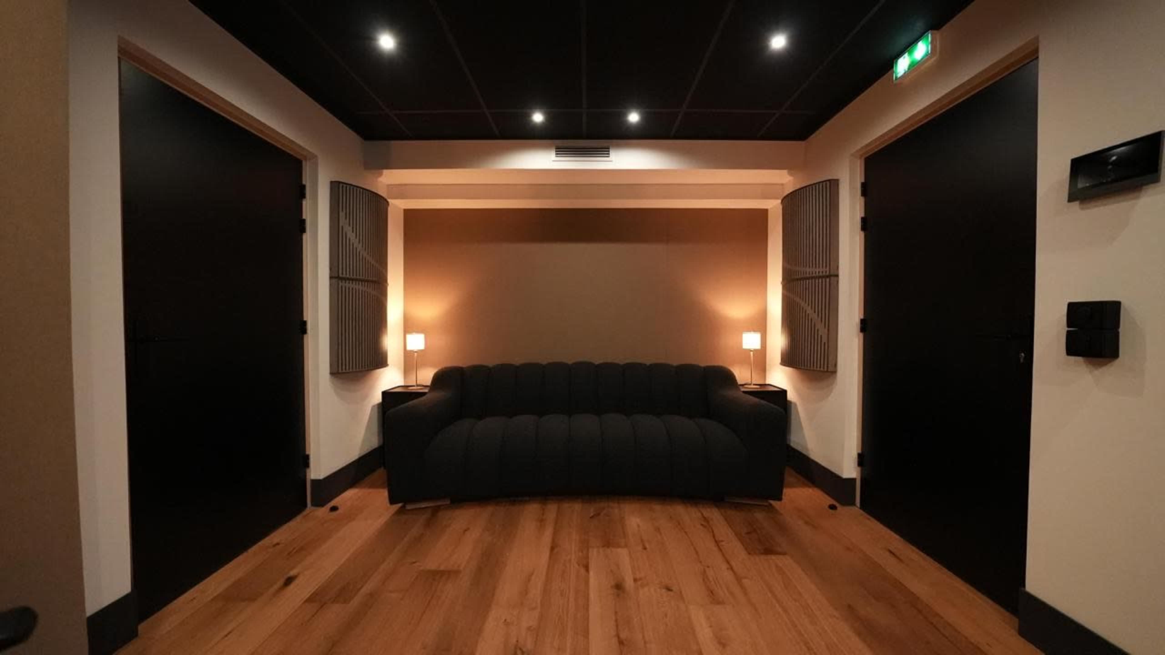 The image shows a clean hallway with a black couch in the center, flanked by two closed black doors, and softly lit by two lamps.