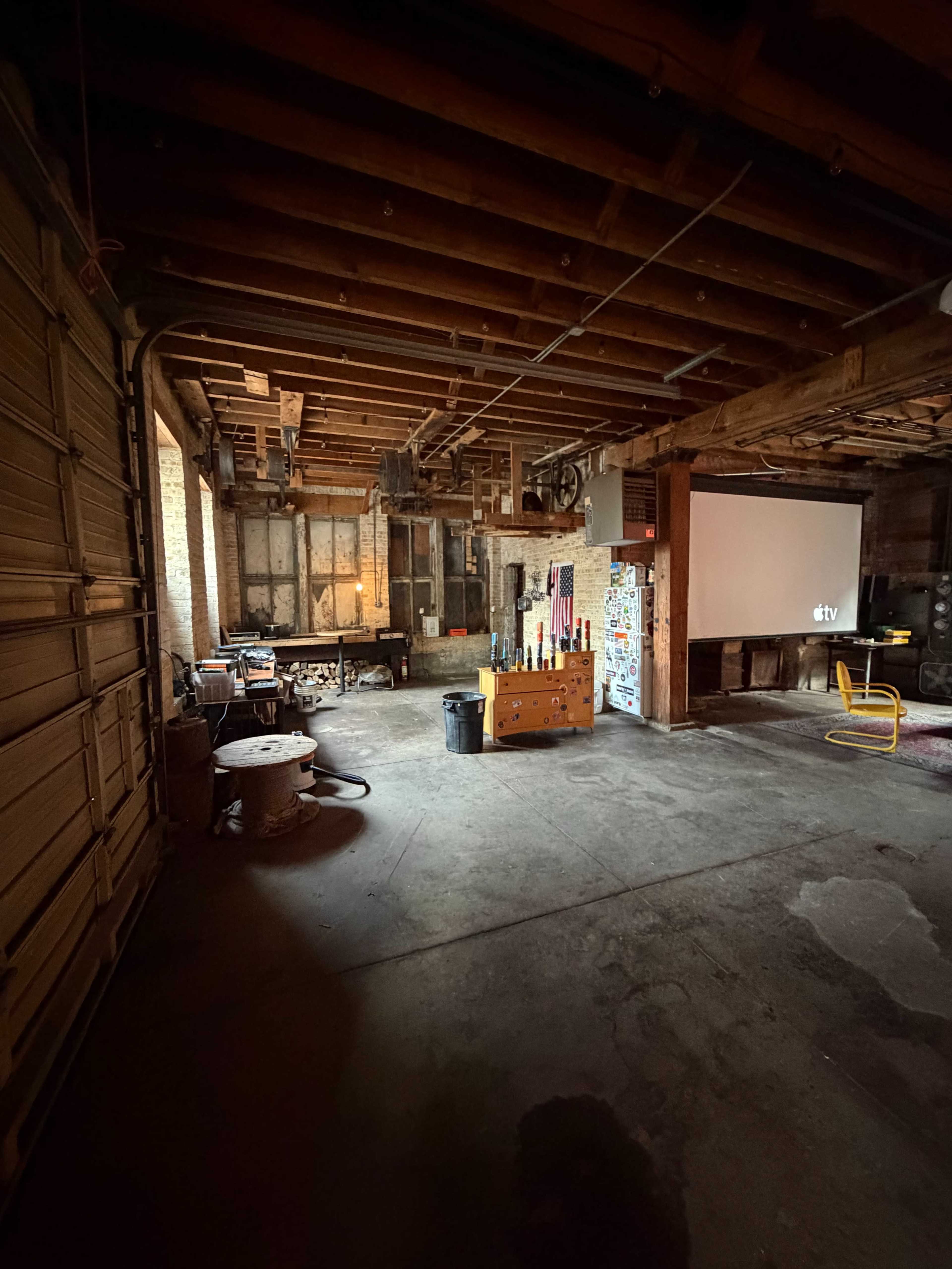 An empty industrial space features exposed wooden beams, a projection screen, and various tools scattered around.
