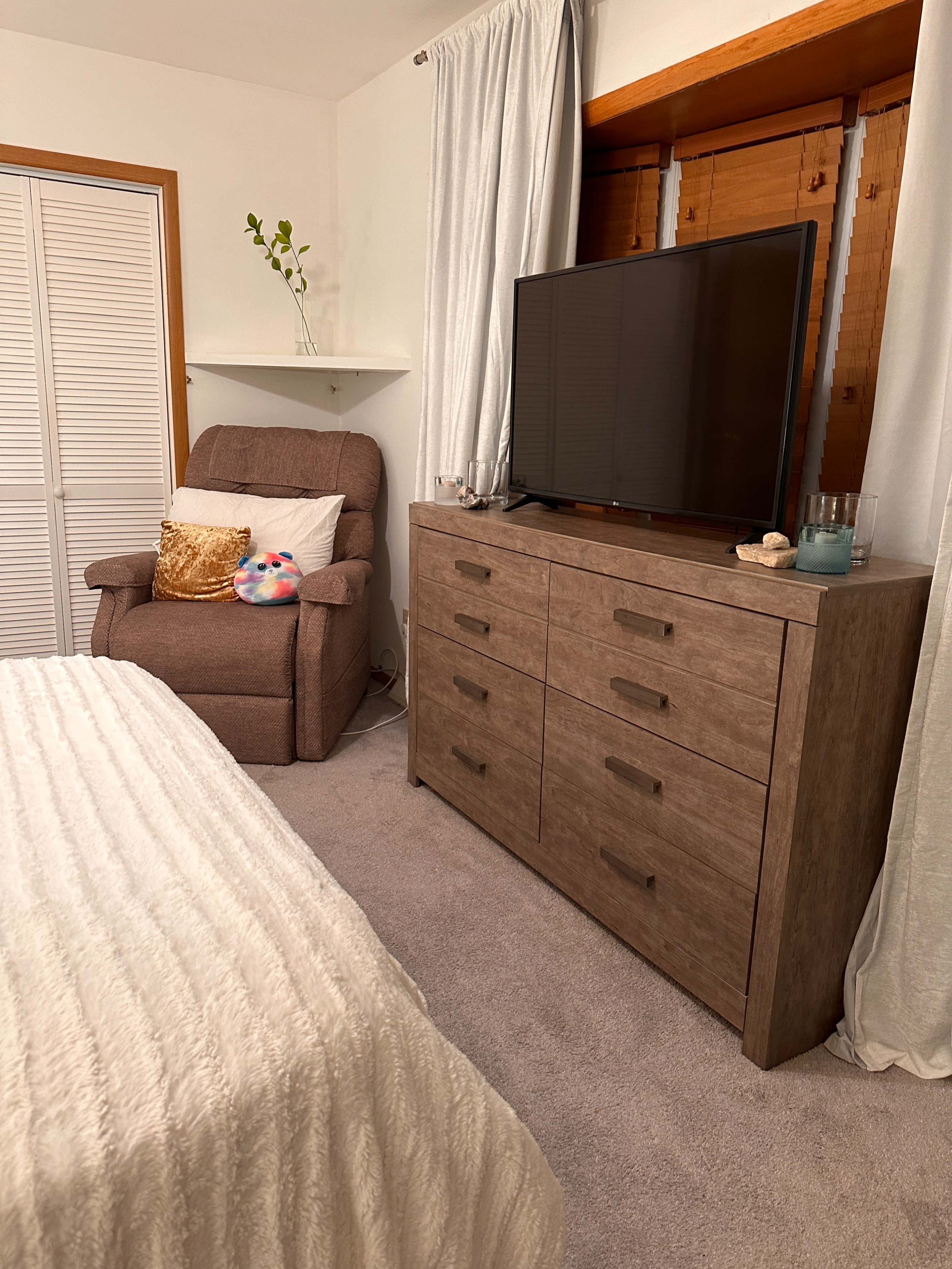 A cozy bedroom featuring a dresser, a television on top, and a recliner chair beside a bed with a textured white blanket.