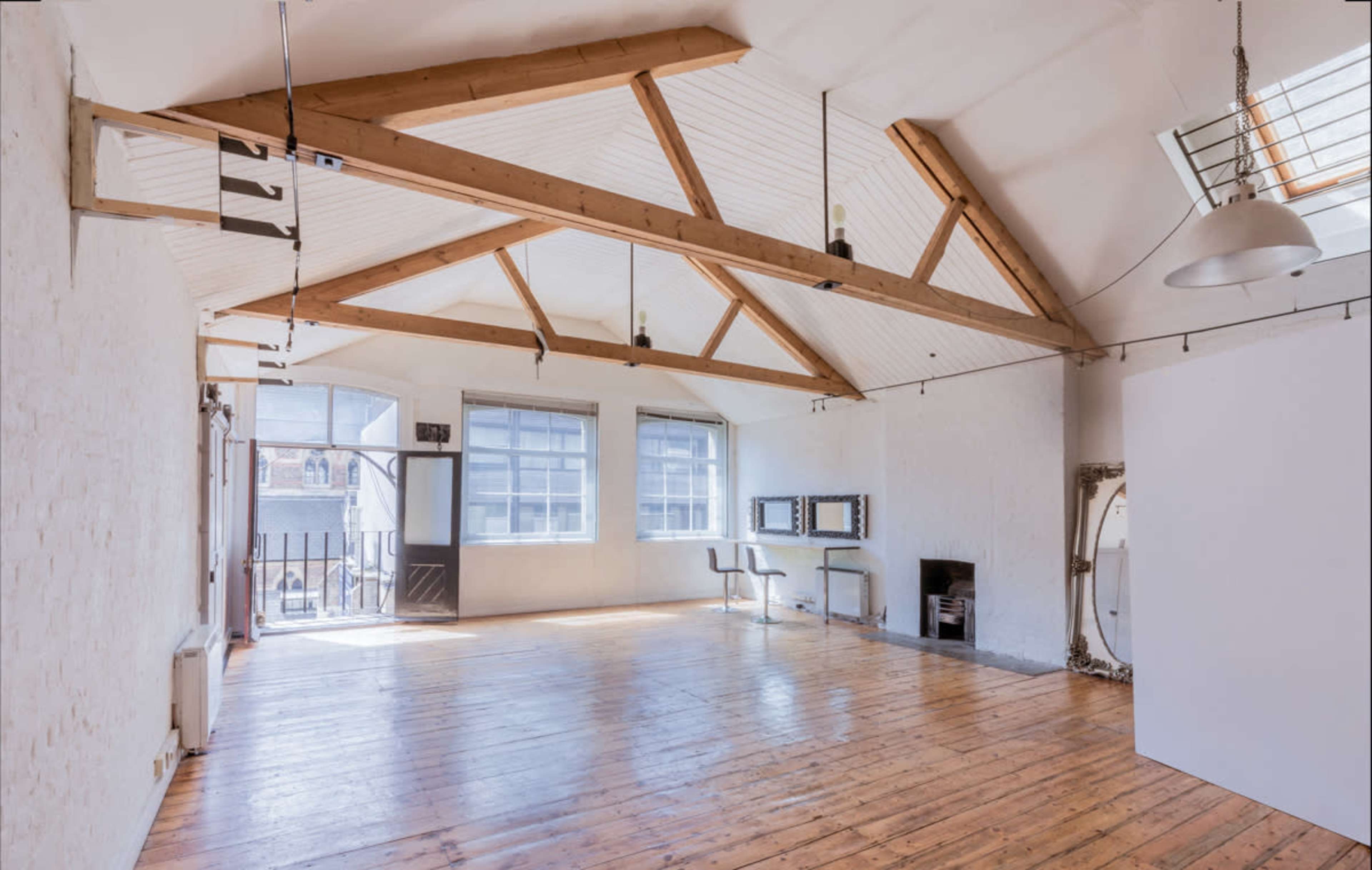 A spacious, well-lit room with wooden floorboards, high ceilings supported by exposed beams, and large windows.