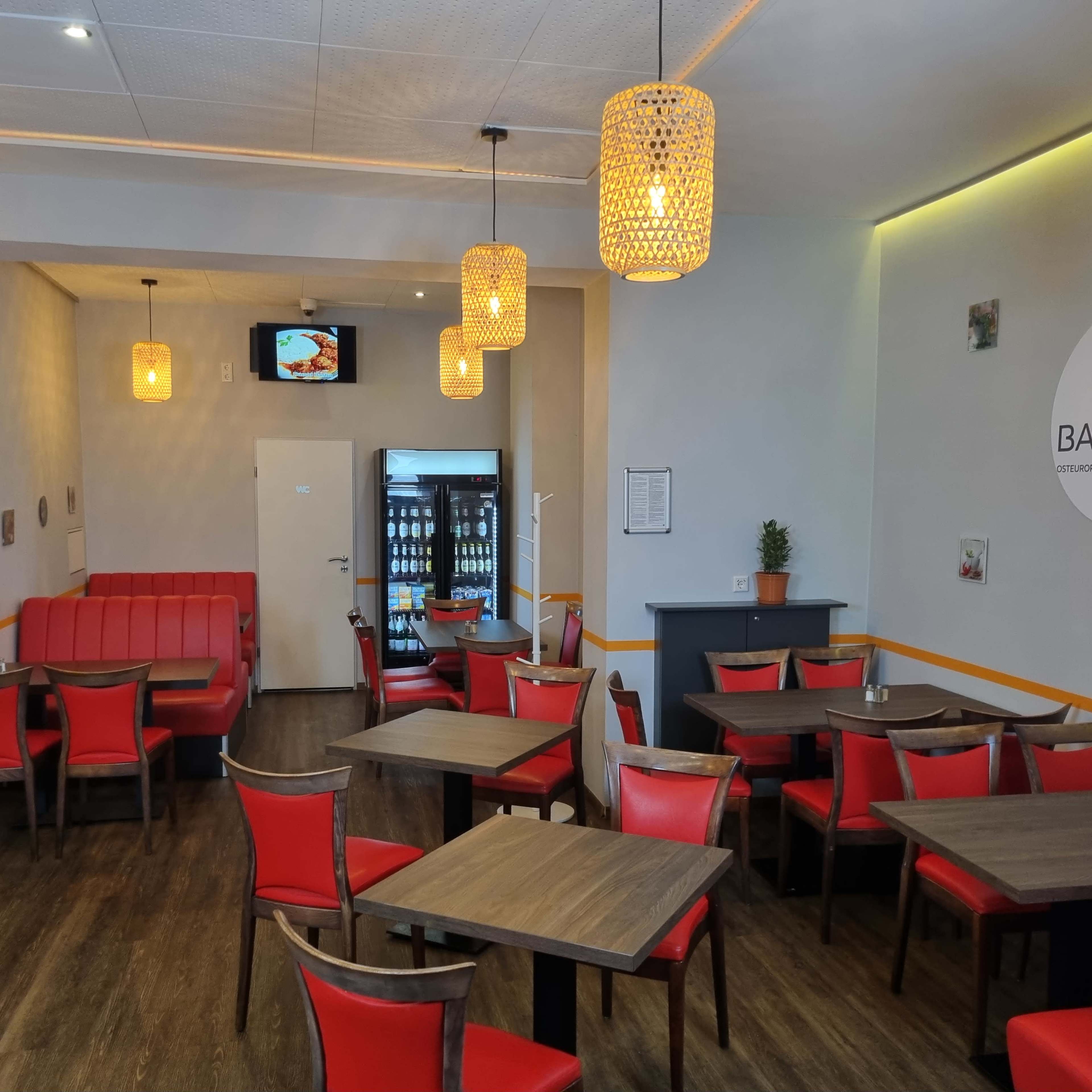 The image shows a modern restaurant interior featuring wooden tables and red chairs, with a seating area and a drink fridge in the background.