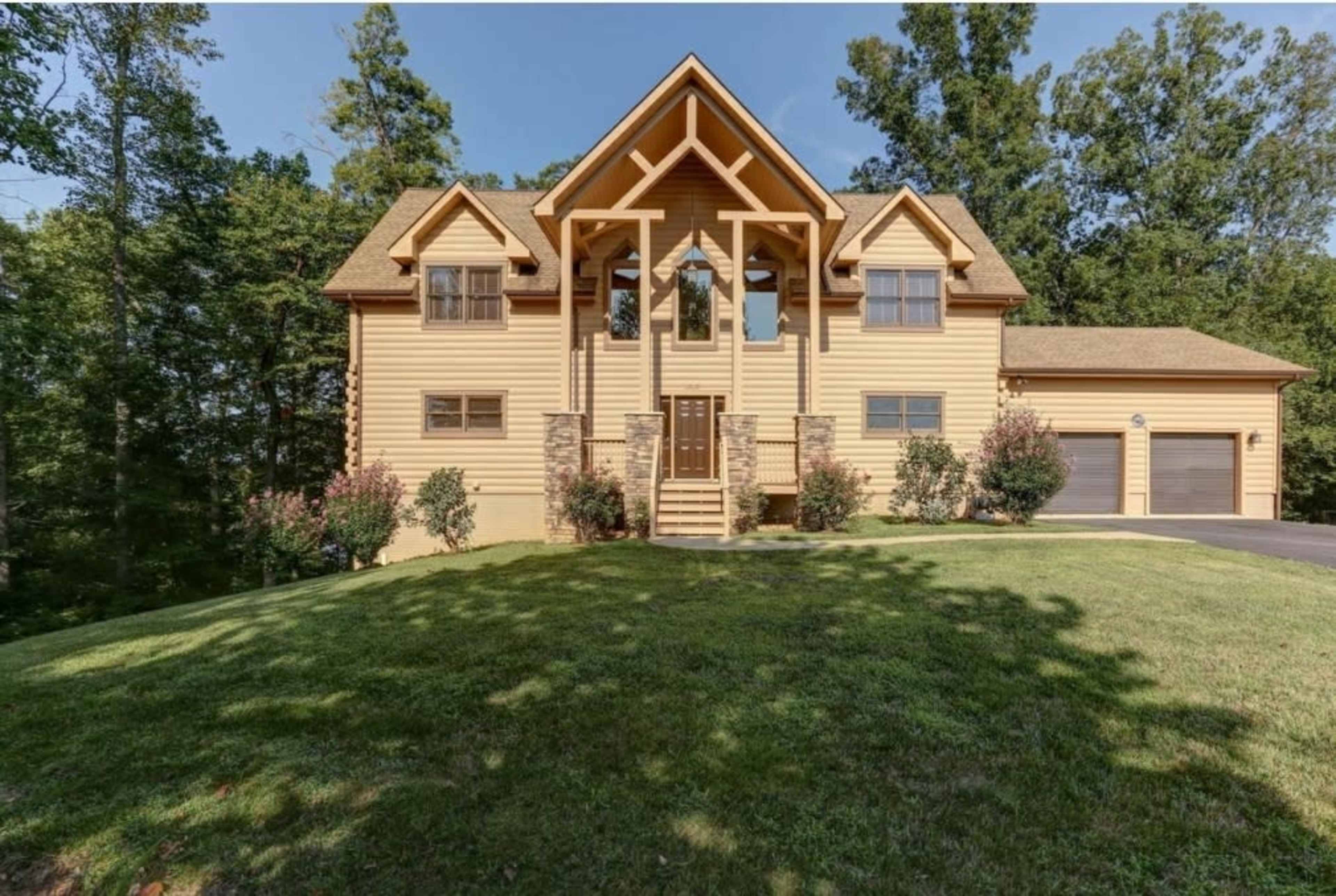 A large, two-story wooden house with a peaked roof, situated on a grassy hill surrounded by trees.