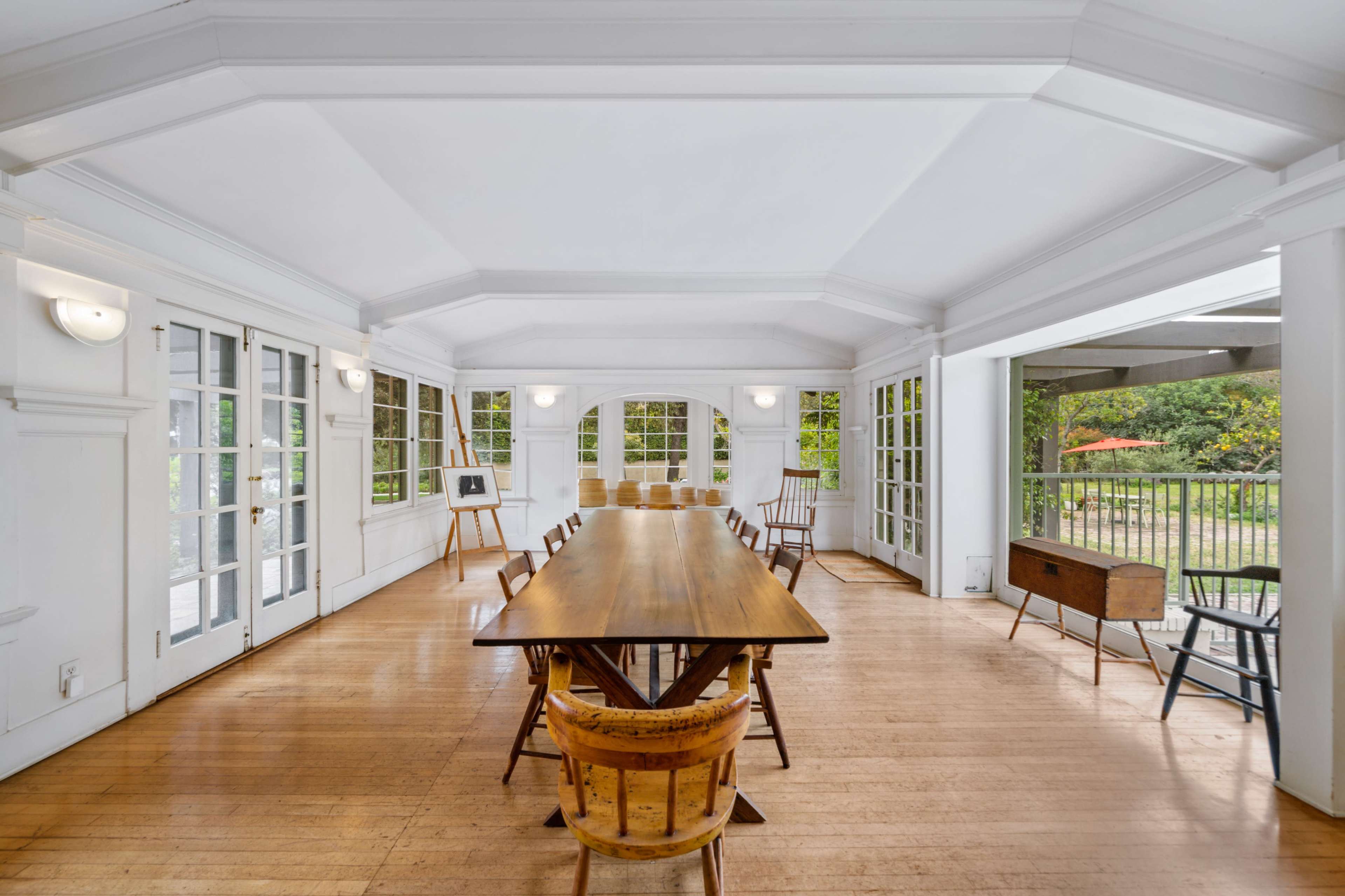A bright, airy room with a long wooden table surrounded by chairs, large windows, and wooden flooring.