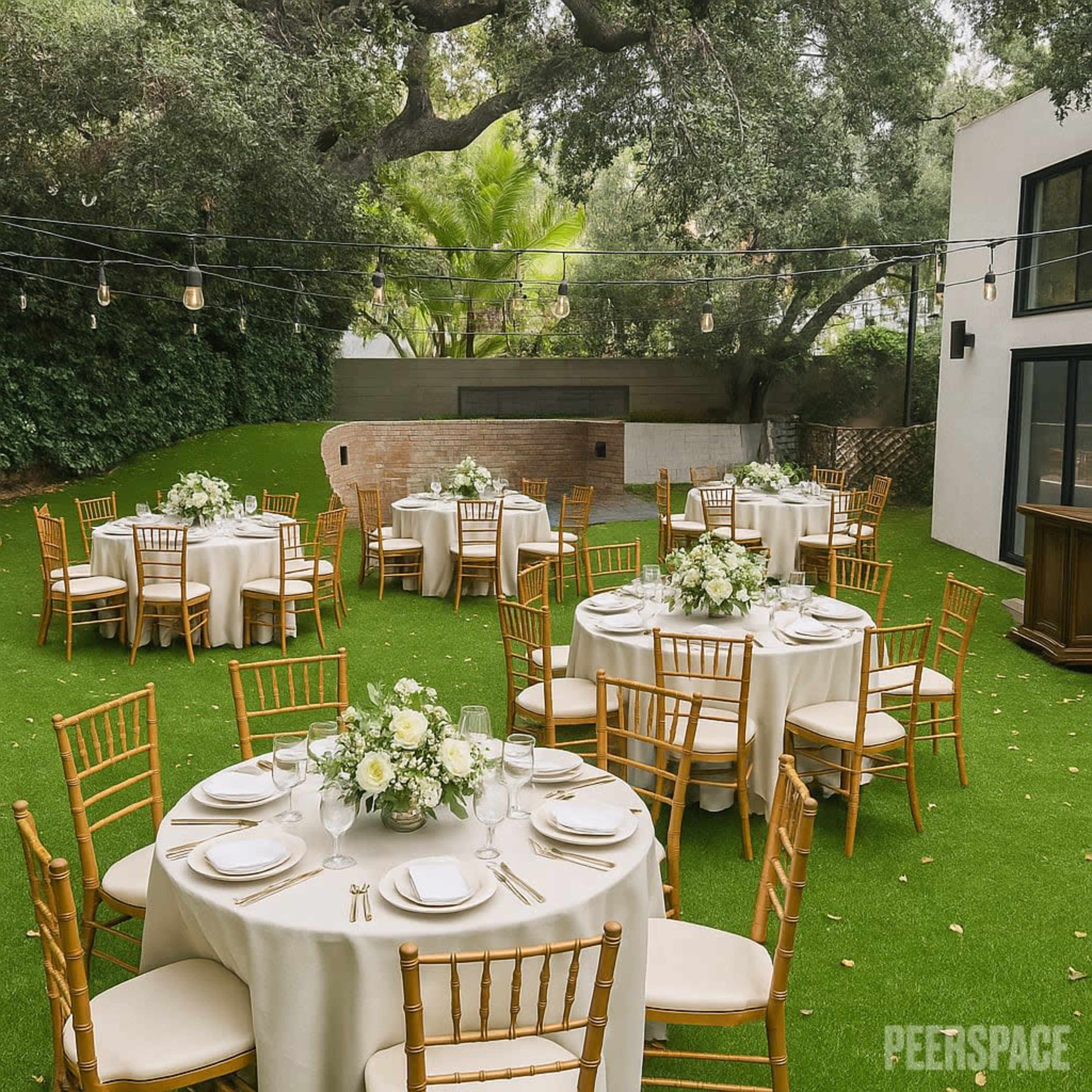 The image shows an outdoor event space with several round tables dressed in cream tablecloths, arranged on a green lawn decorated with white floral centerpieces and surrounded by trees and string lights.