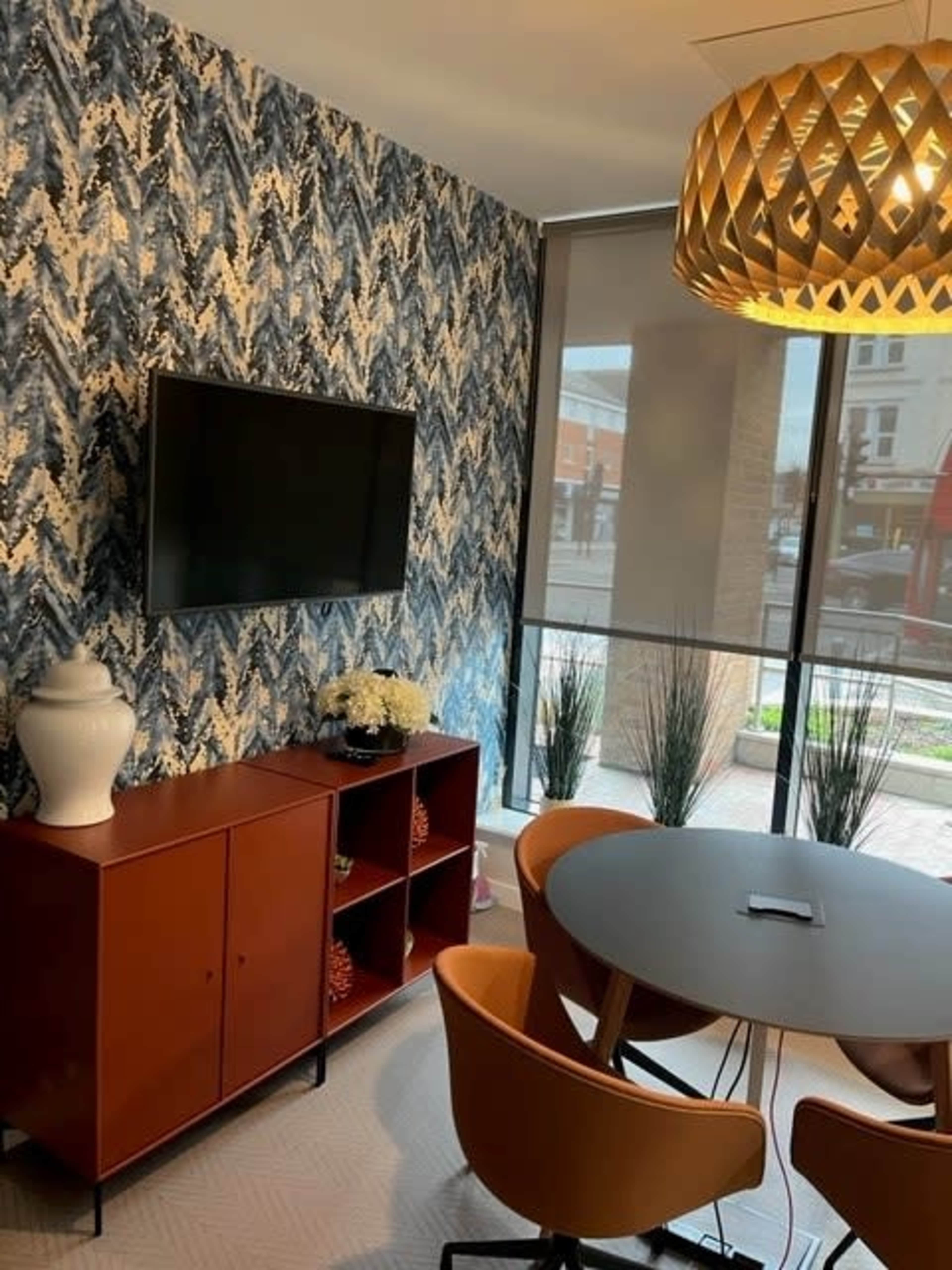 A modern indoor space features a round table with orange chairs, a flat-screen TV on a decorative wall, and a cabinet beside large windows.