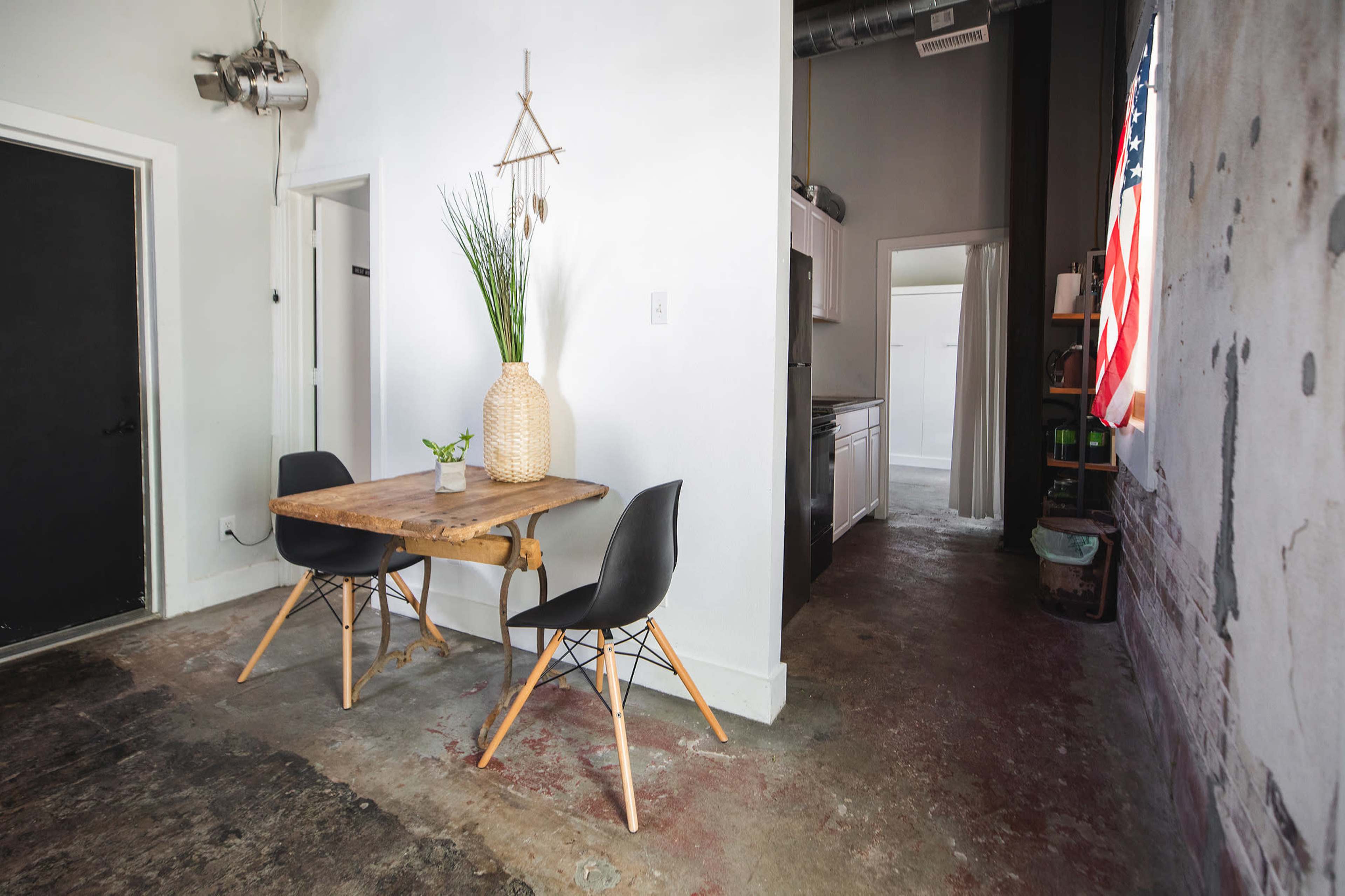 A wooden table with two black chairs is set against a wall in a room featuring a visible kitchen and an American flag hanging nearby.