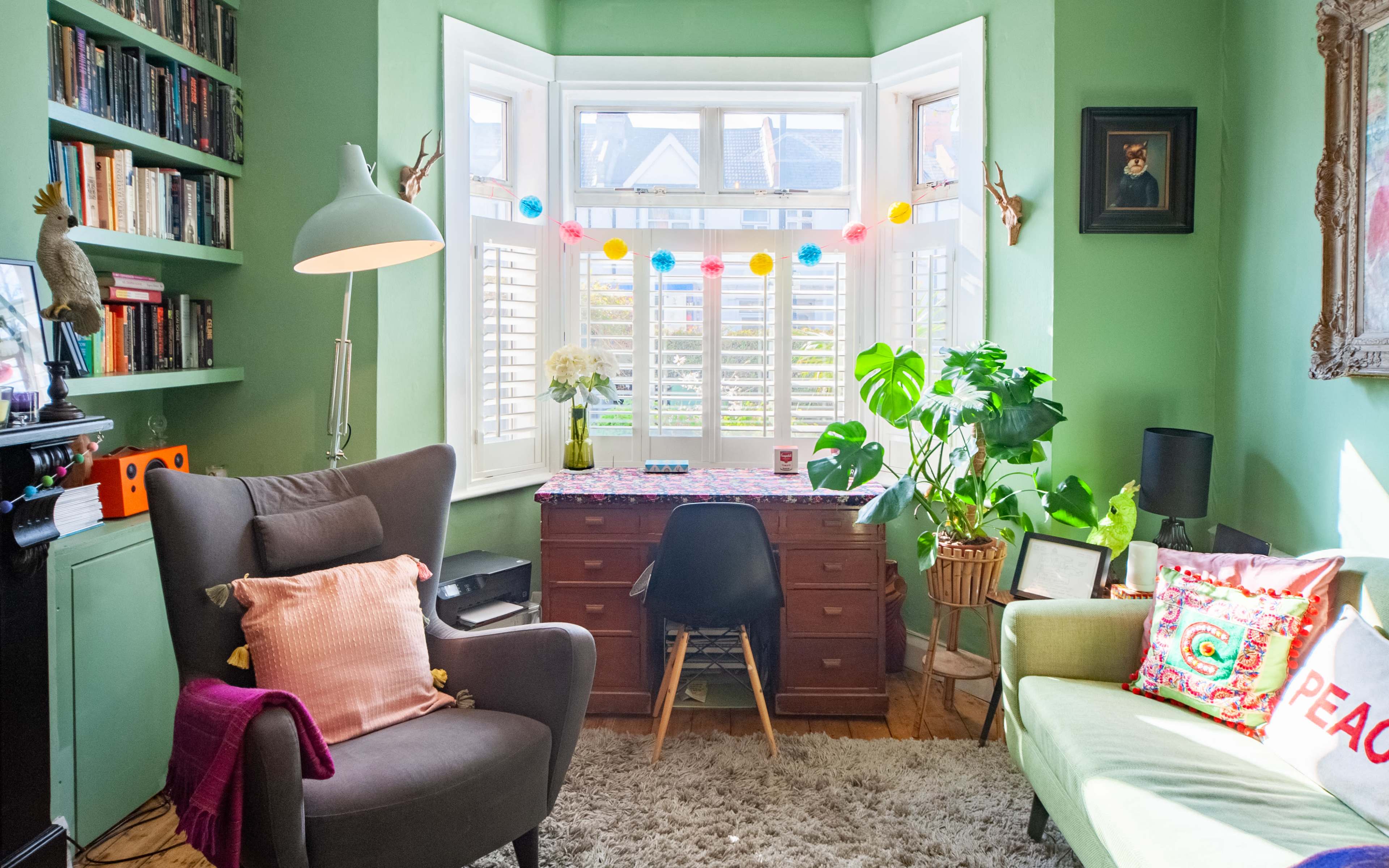 A cozy study features a wooden desk in front of a bay window, surrounded by plants, bookshelves, and comfortable seating.