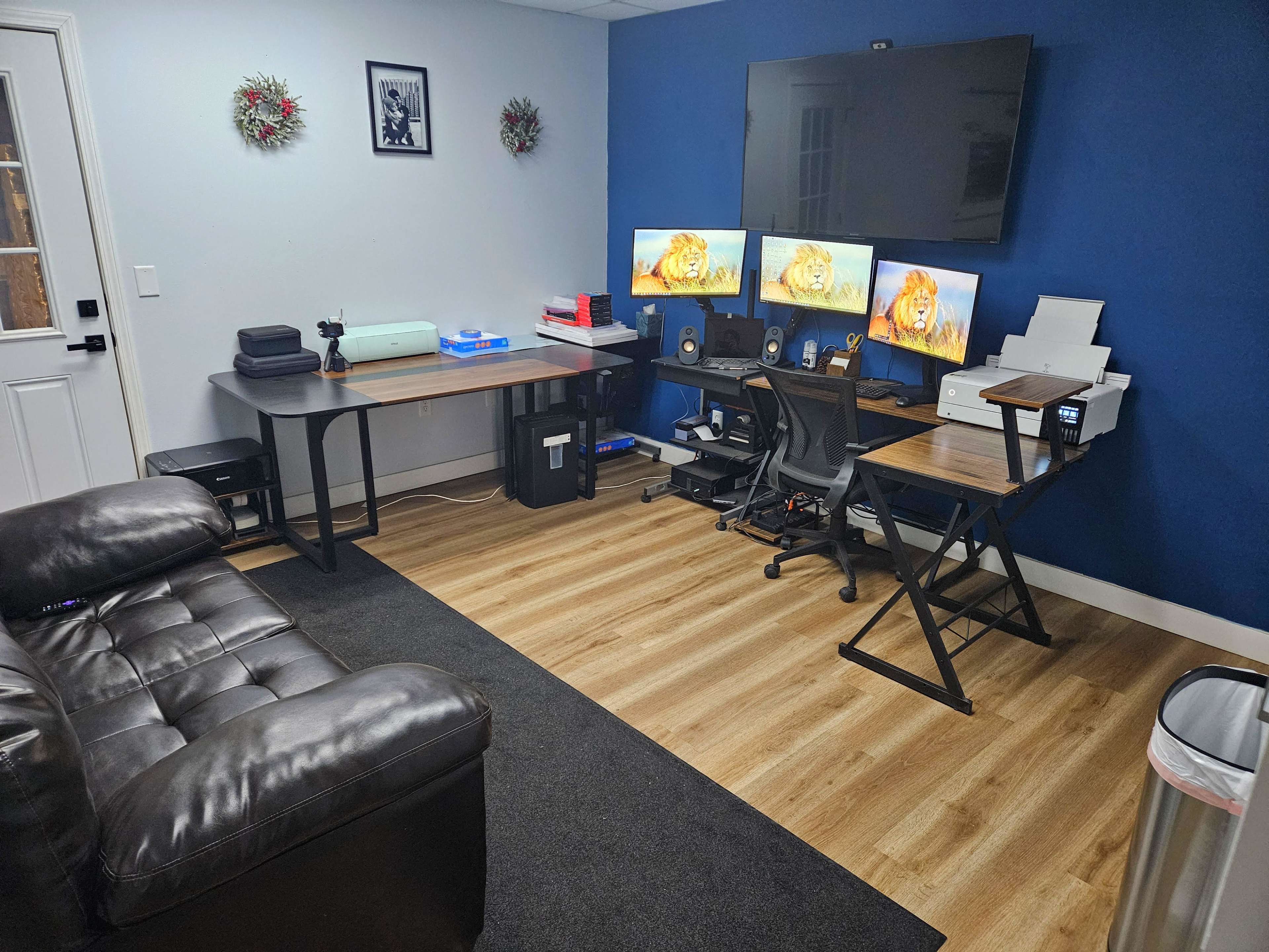 The image shows an office space featuring two desks with multiple computer monitors, a couch, and a wall-mounted television against a blue wall.