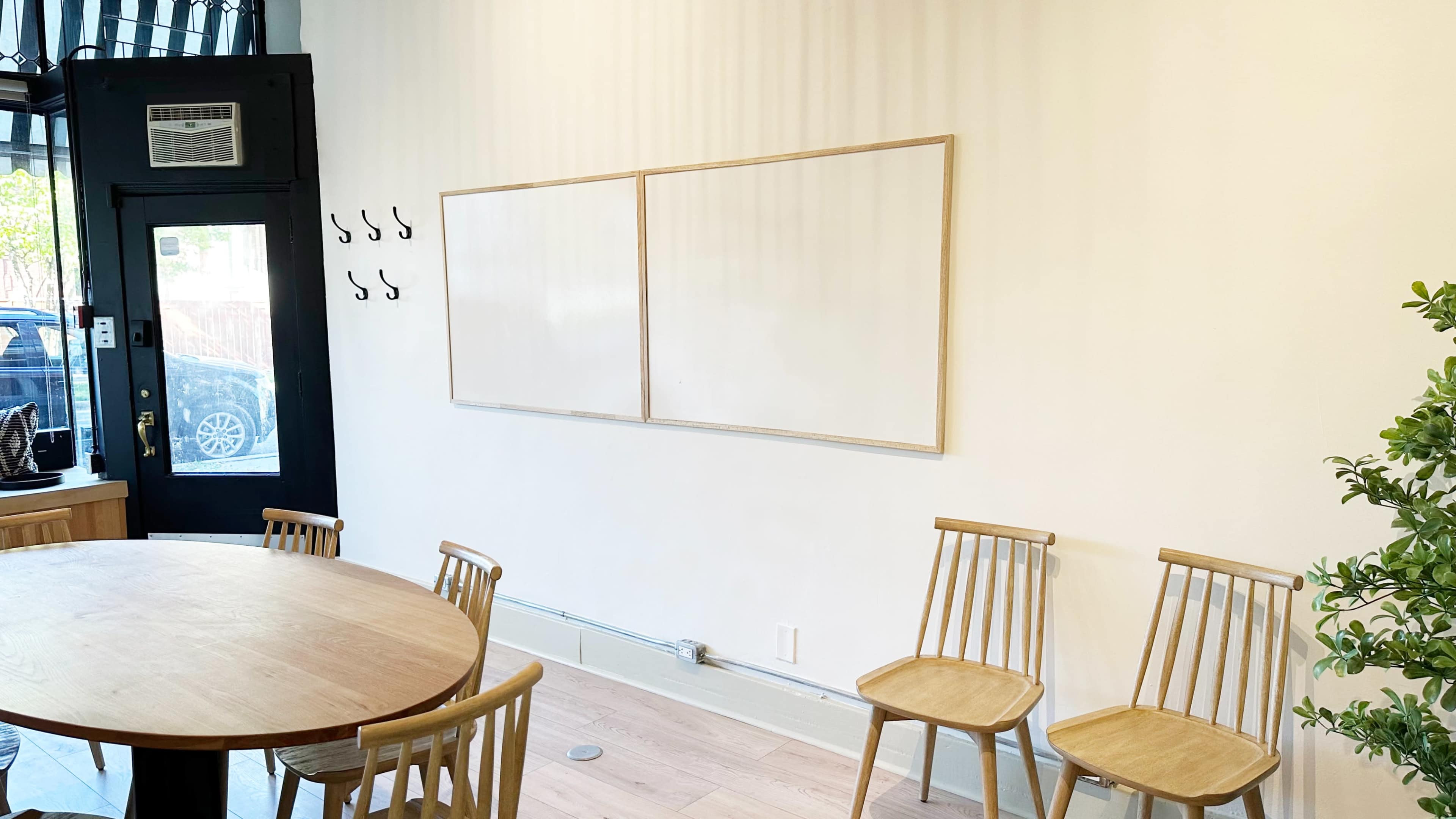 The image shows a minimalistic room featuring a round wooden table surrounded by five wooden chairs, with a blank wall that has two framed whiteboards and several hooks on the left side.