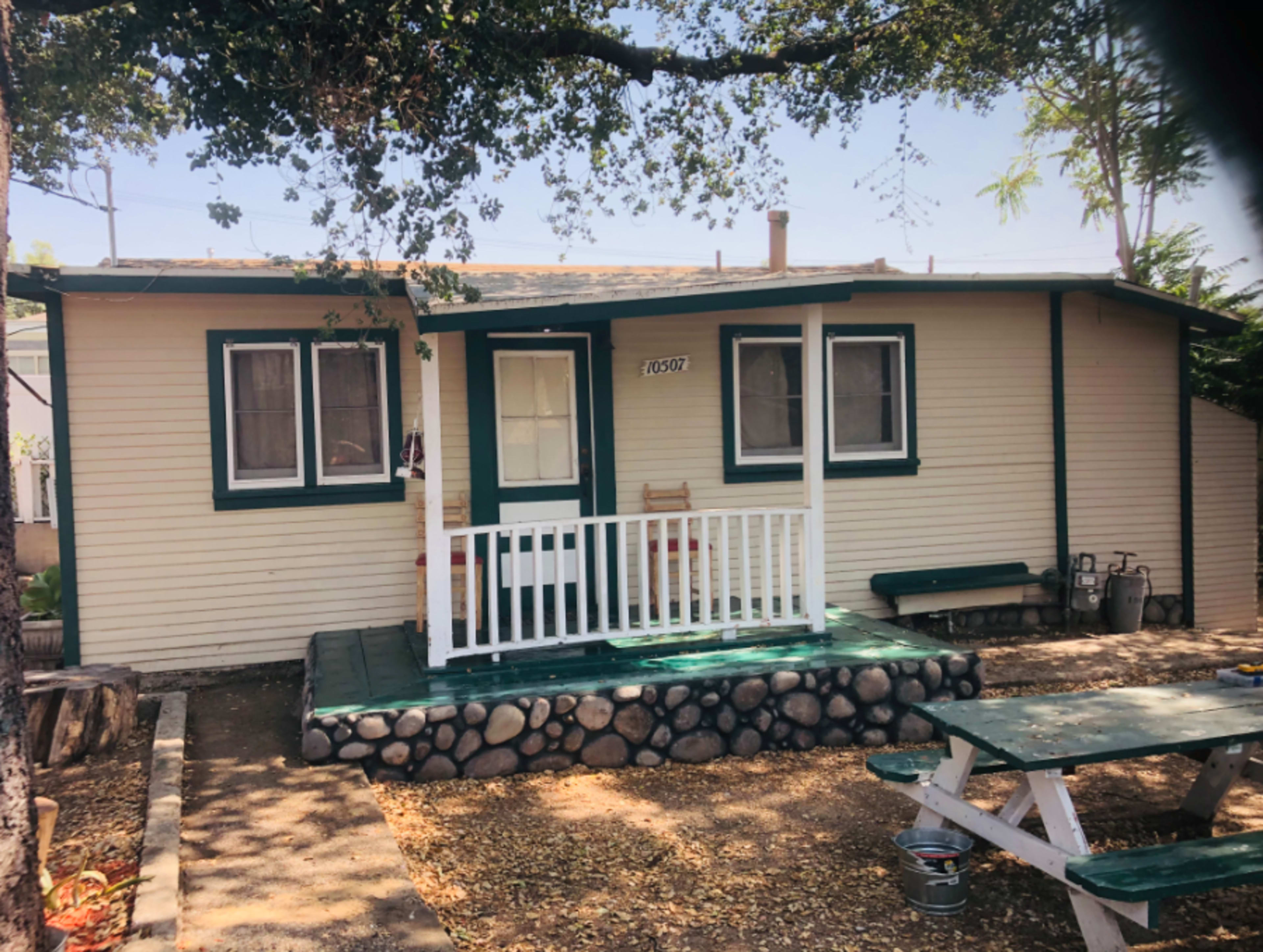 A small, single-story house with a porch and green trim, surrounded by a gravel yard and a picnic table.