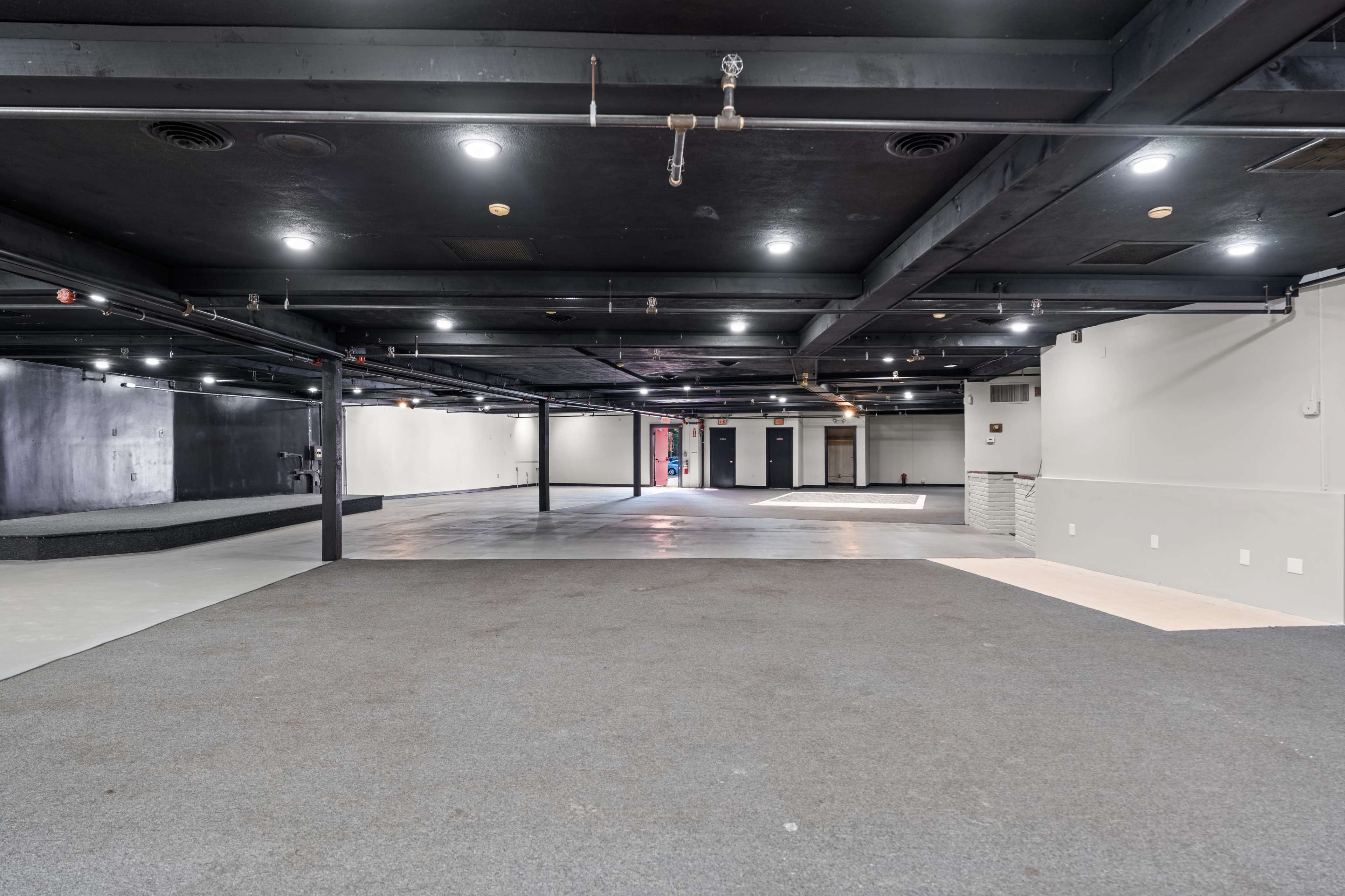 The image shows a large, sparsely furnished indoor space with dark walls, multiple ceiling lights, and grey carpeting.