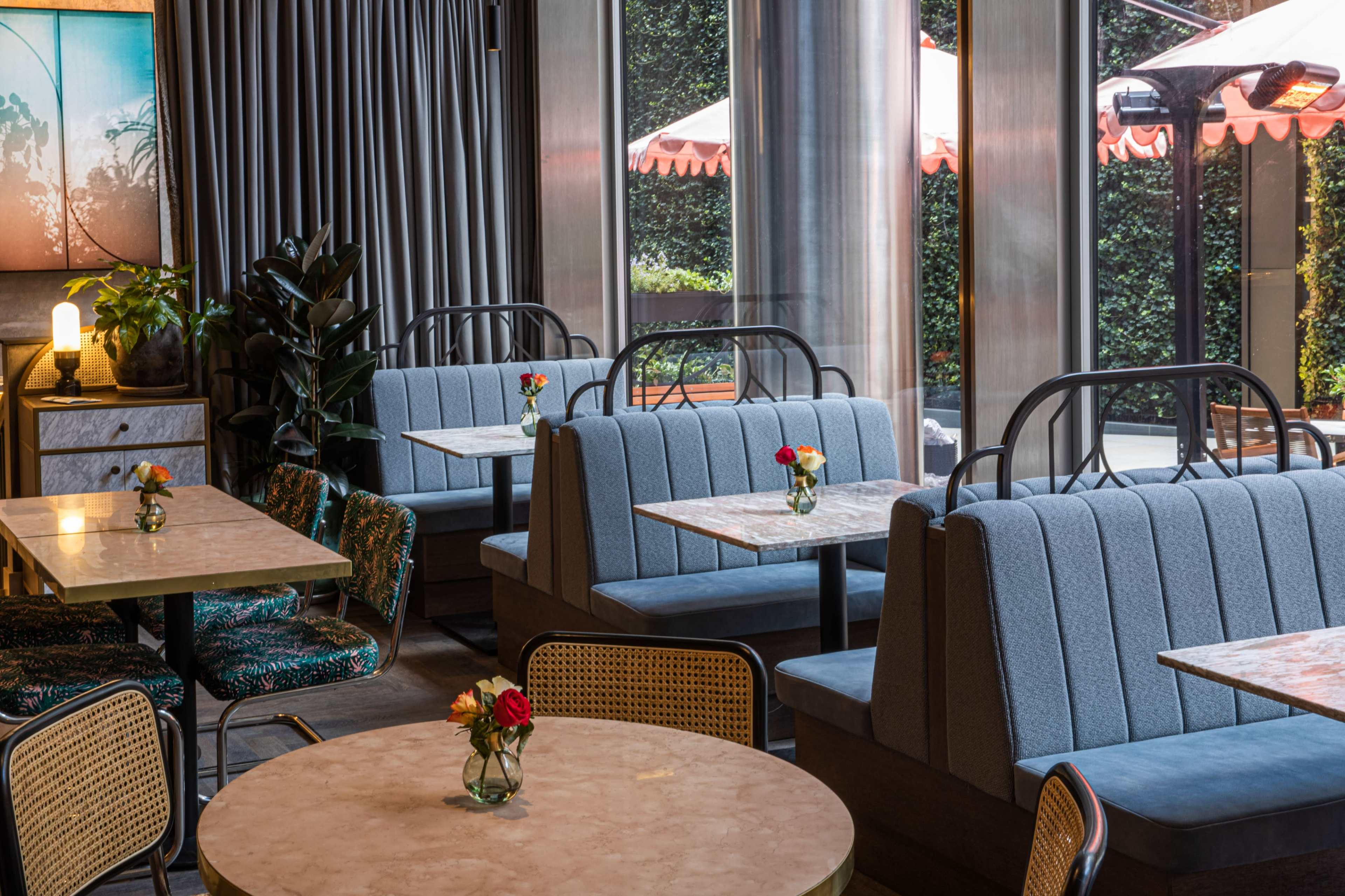 The image shows a modern cafe interior with blue upholstered booths, round tables, and small flower vases, surrounded by large windows that let in natural light.