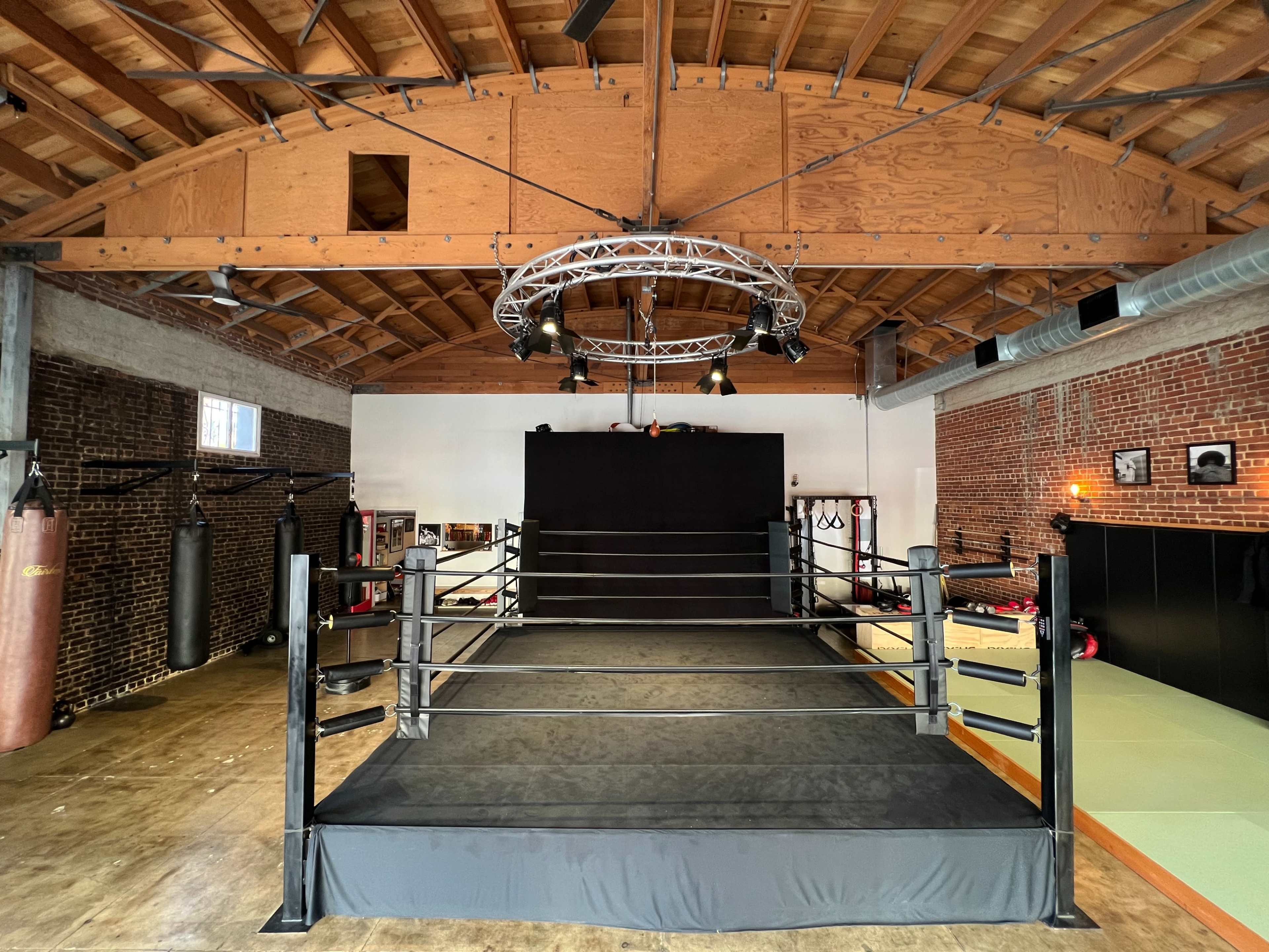 An indoor boxing gym featuring a central boxing ring, hanging punch bags, and a black backdrop.