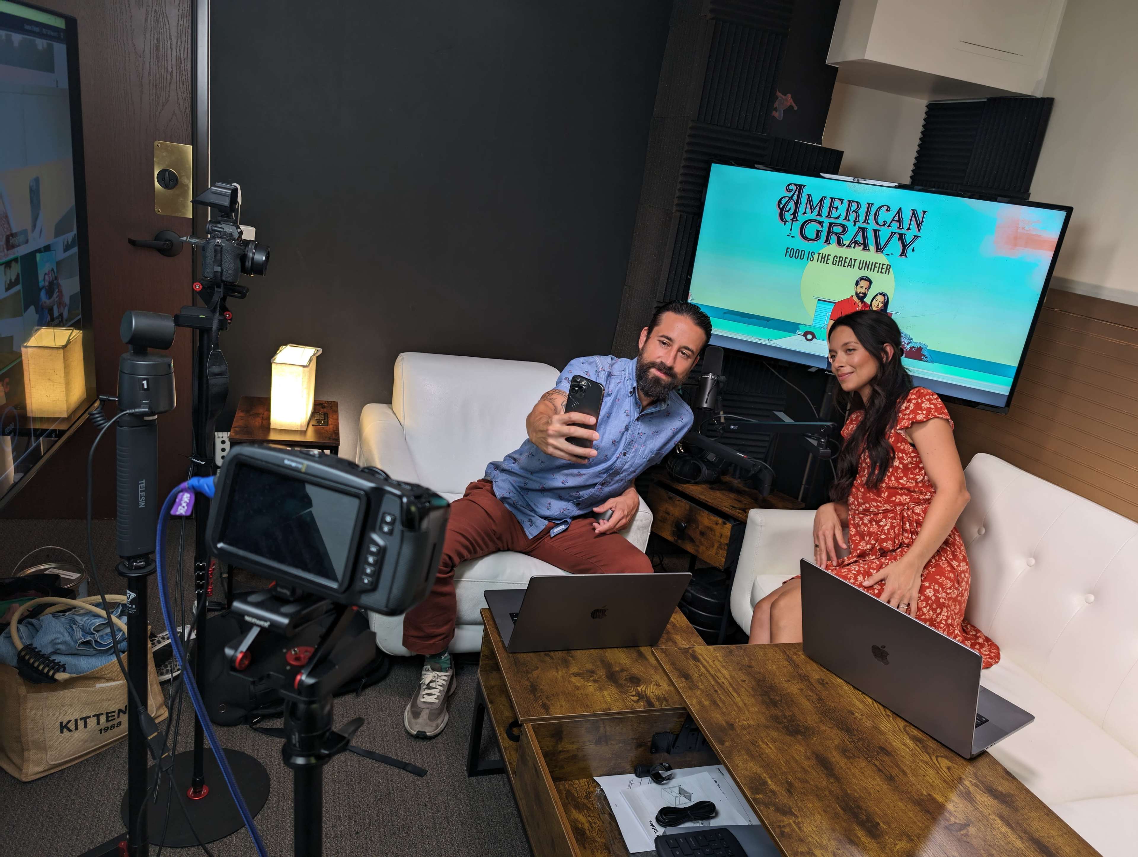 A man and a woman sit in a studio equipped for filming, with cameras and laptops set up in front of them while they take a selfie.
