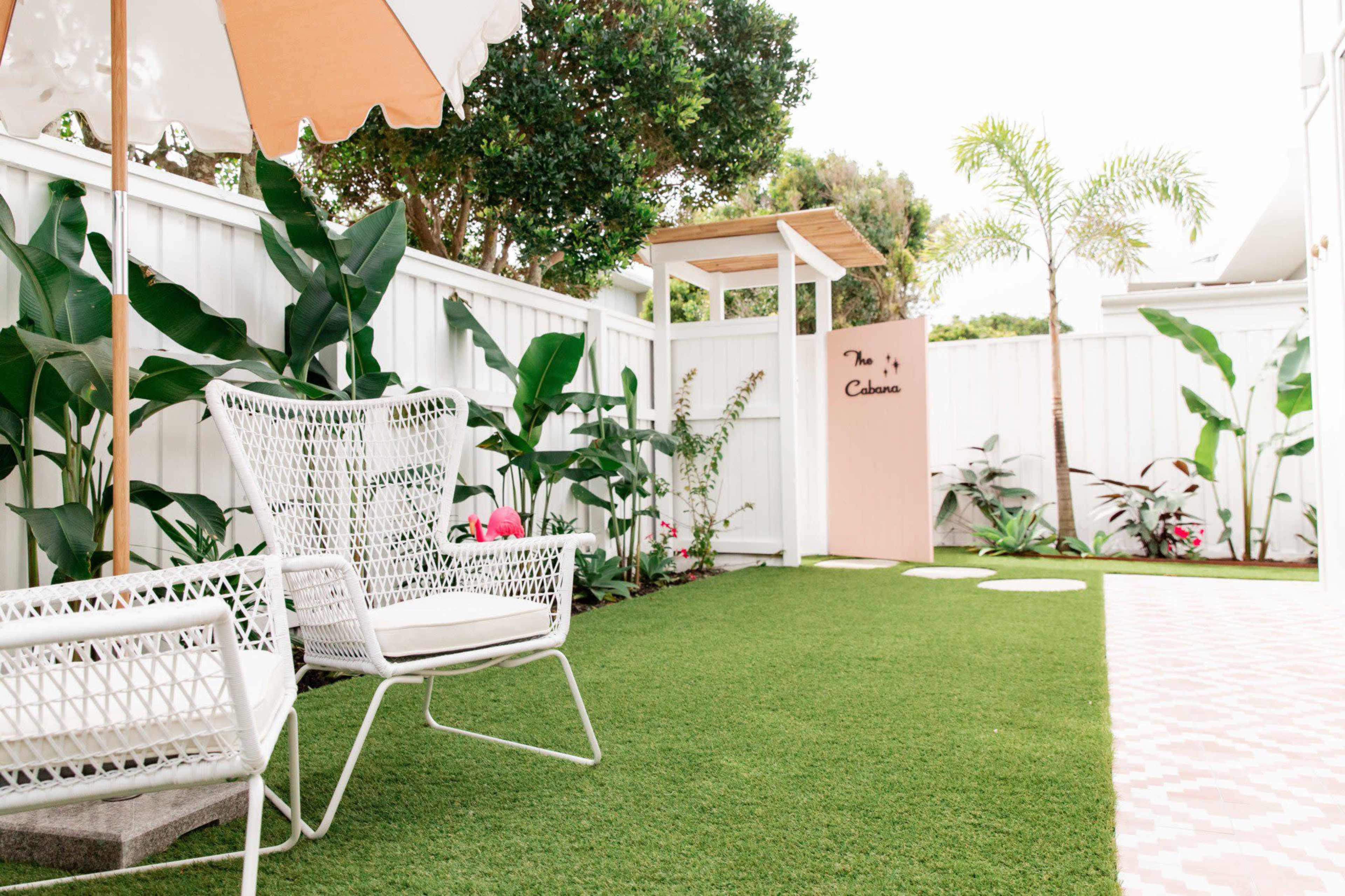 A landscaped outdoor space features white wicker chairs, artificial grass, tall green plants, a pink wall structure, and a shaded umbrella.