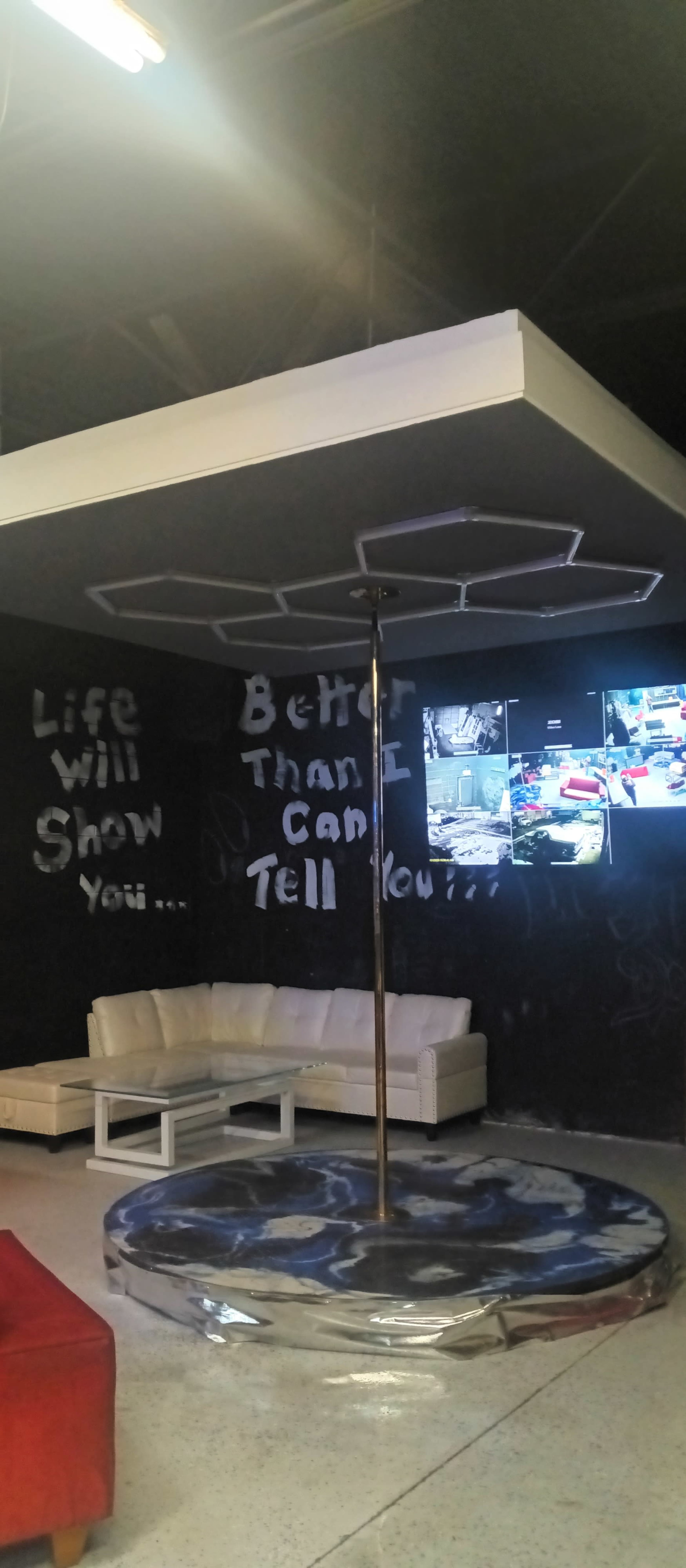 An empty interior space featuring a pole in the center, surrounded by a white couch and screens displaying video footage on a dark wall.