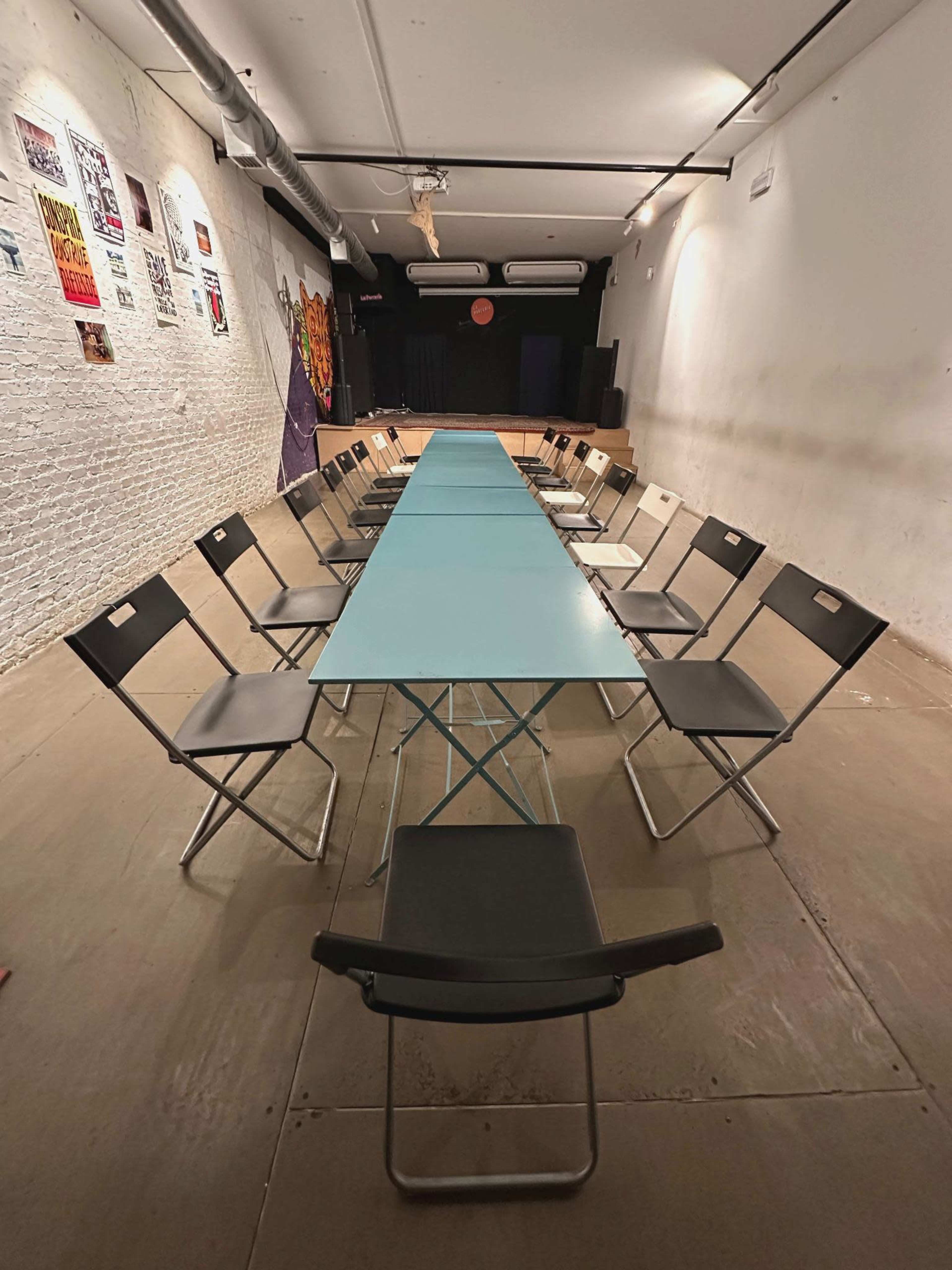 A long, blue table with black folding chairs on either side is set up in a sparsely decorated room with exposed brick walls.