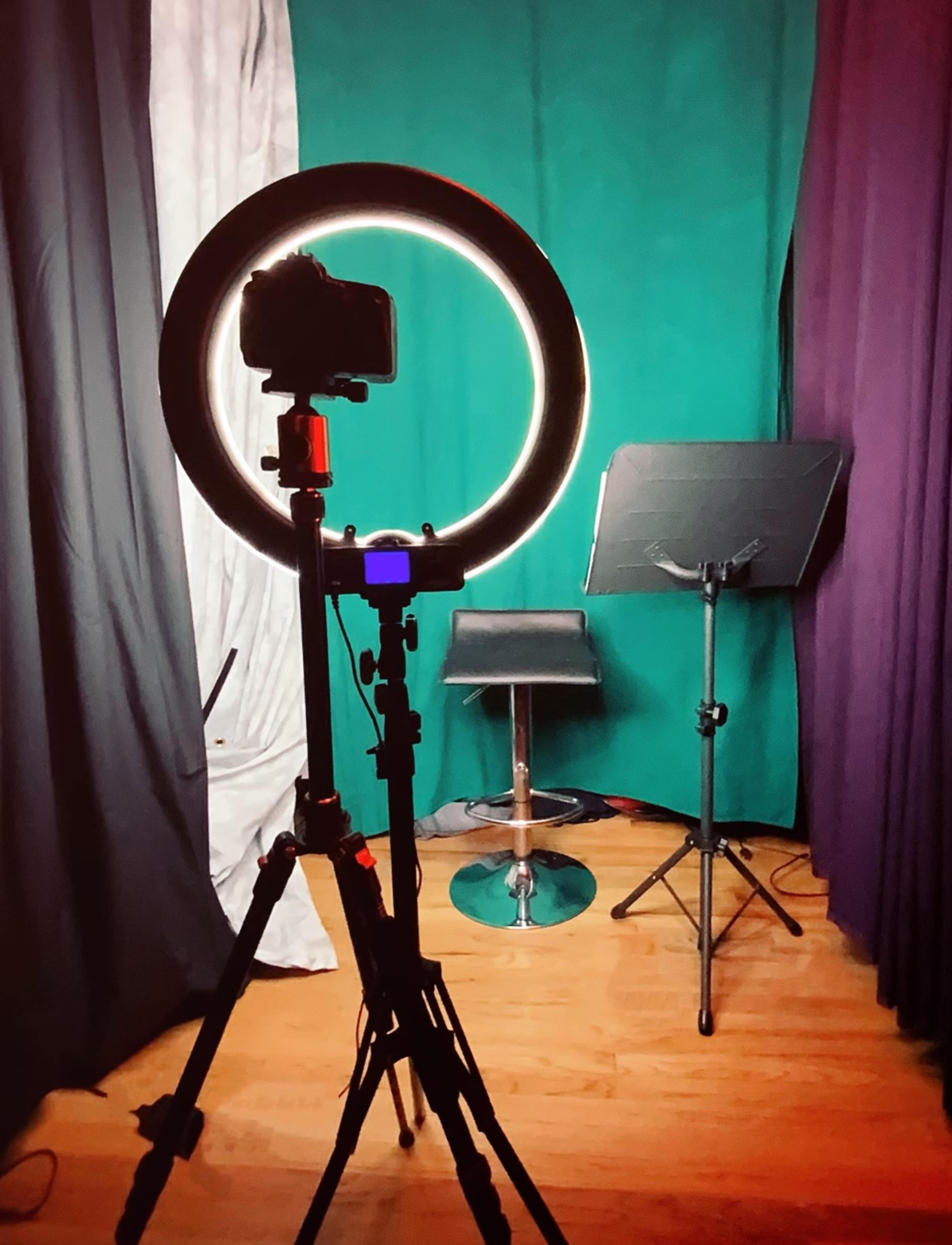A photography setup with a camera mounted on a tripod in front of a circular light, a music stand, and a bar stool against a green backdrop.