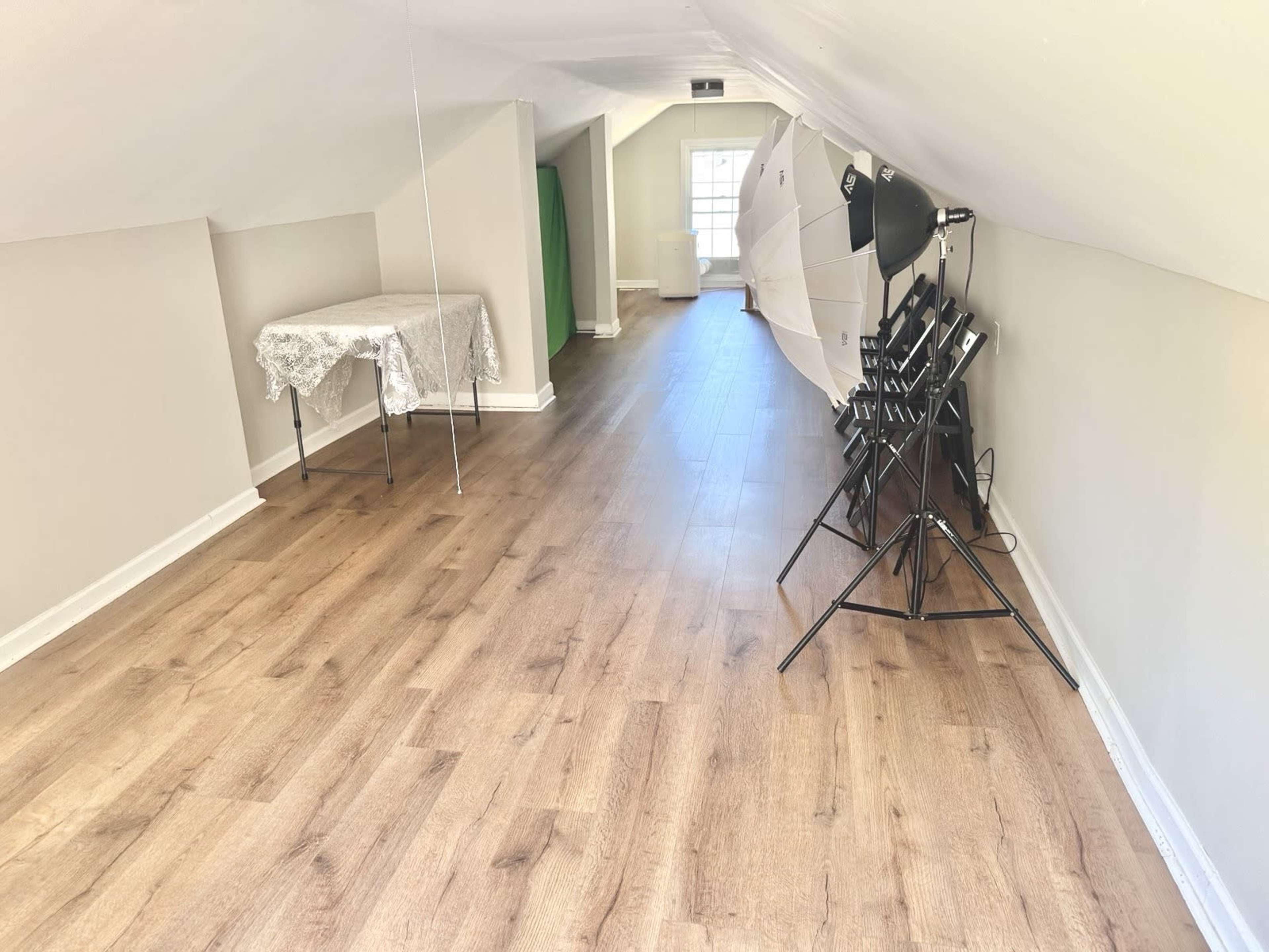 The image shows a well-lit attic space with wooden flooring, a table covered with a lace tablecloth, a green backdrop, and a photography lighting setup on one side.