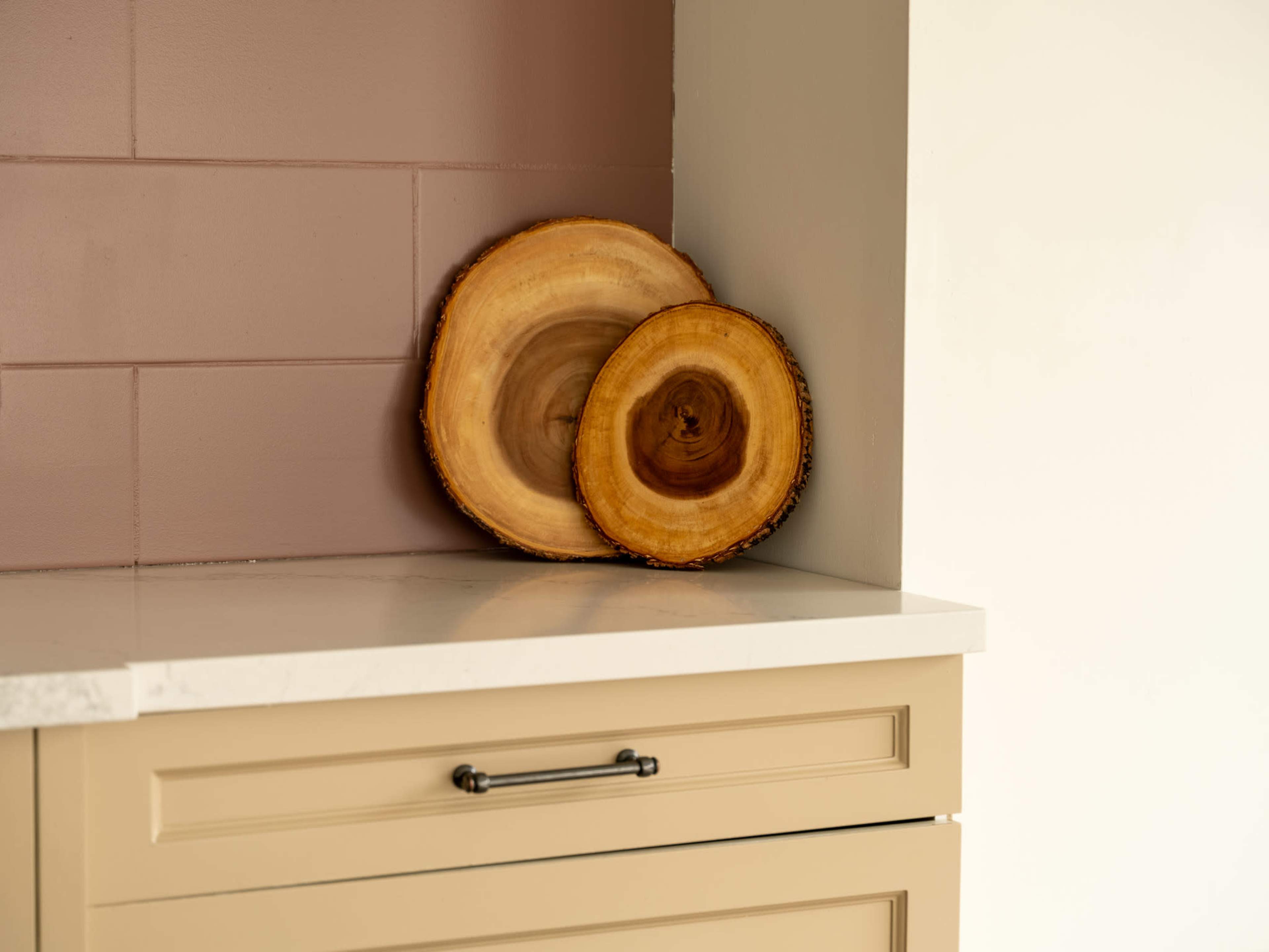 Two wooden slices are displayed against a muted pink wall on a counter with light-colored cabinetry.