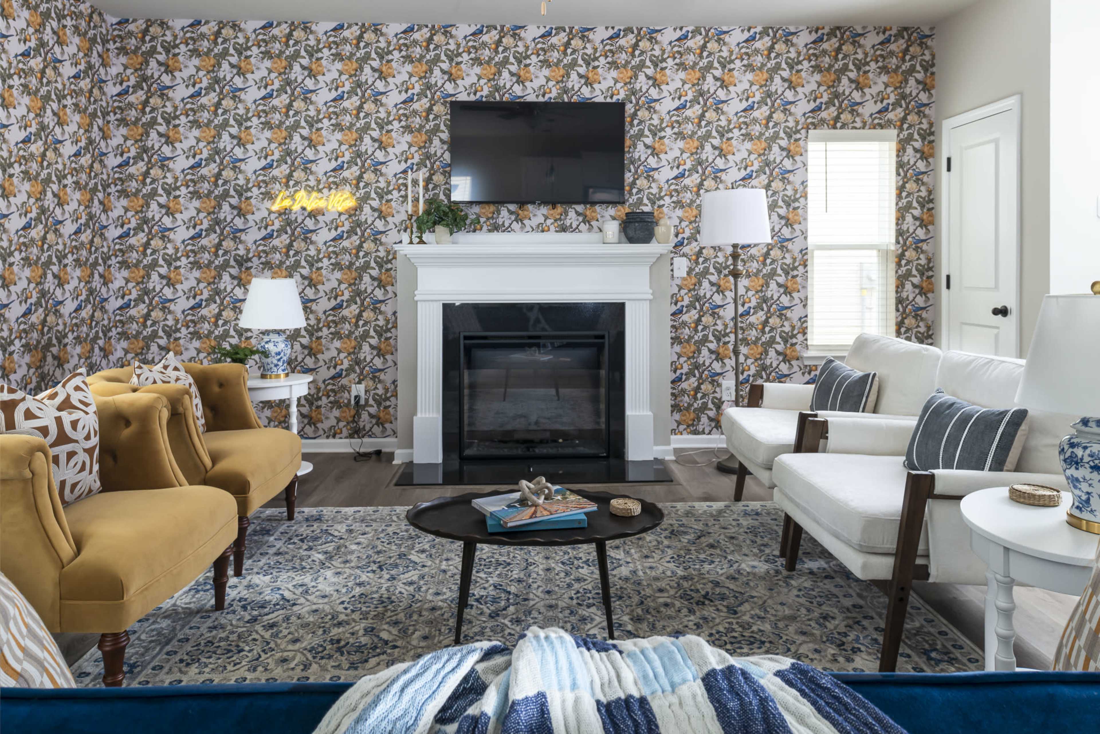 The living room features a floral-patterned wallpaper, a white fireplace with a television mounted above it, two armchairs, a white sofa, and a round coffee table.