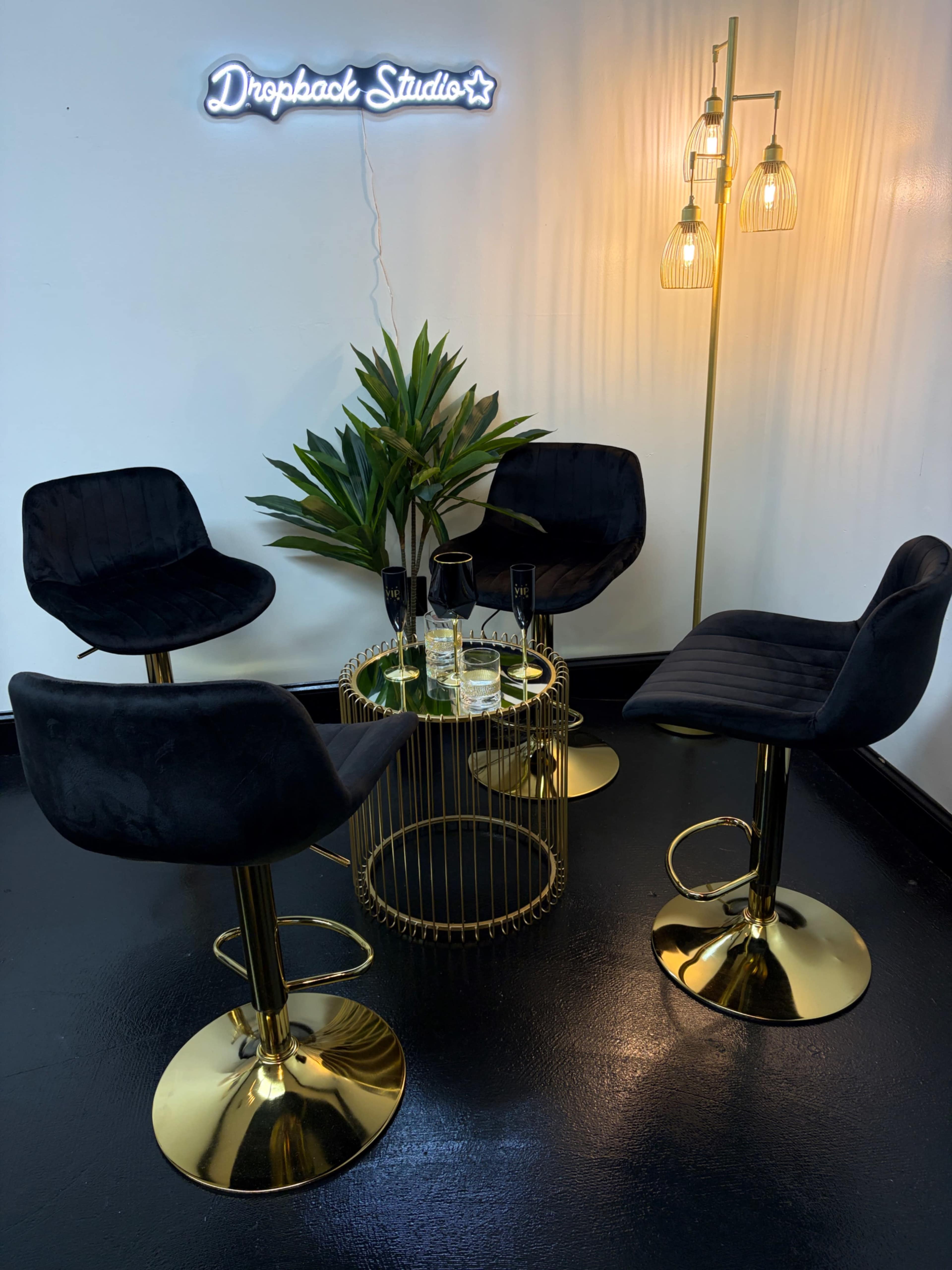 A small gathering area features four black high chairs around a gold circular table, accompanied by a potted plant and a standing lamp against a white wall with a neon sign reading "Dropback Studio."