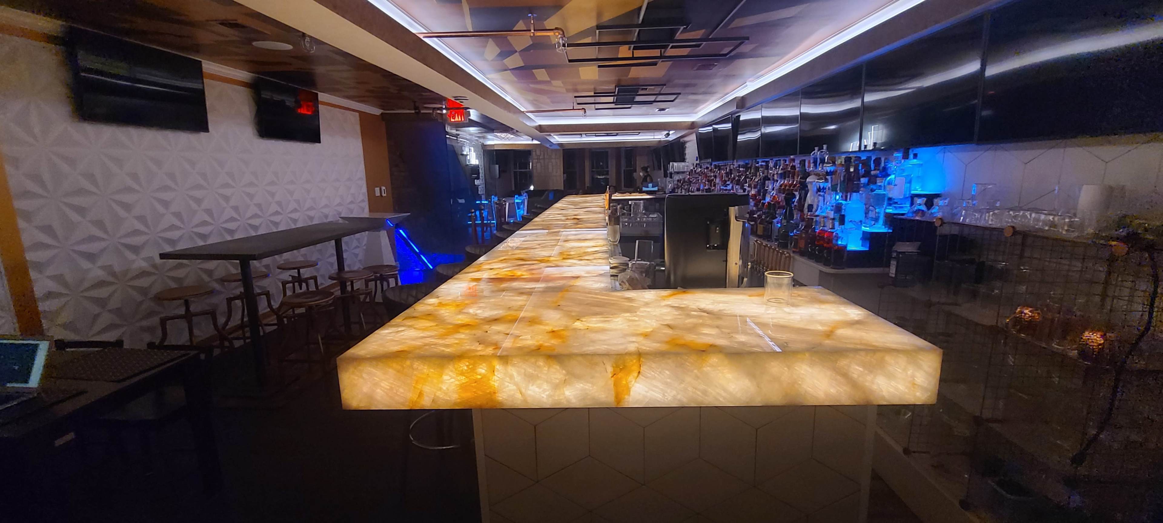 The image shows a modern bar with a brightly lit onyx countertop, sleek shelves stocked with various drinks, and illuminated walls.