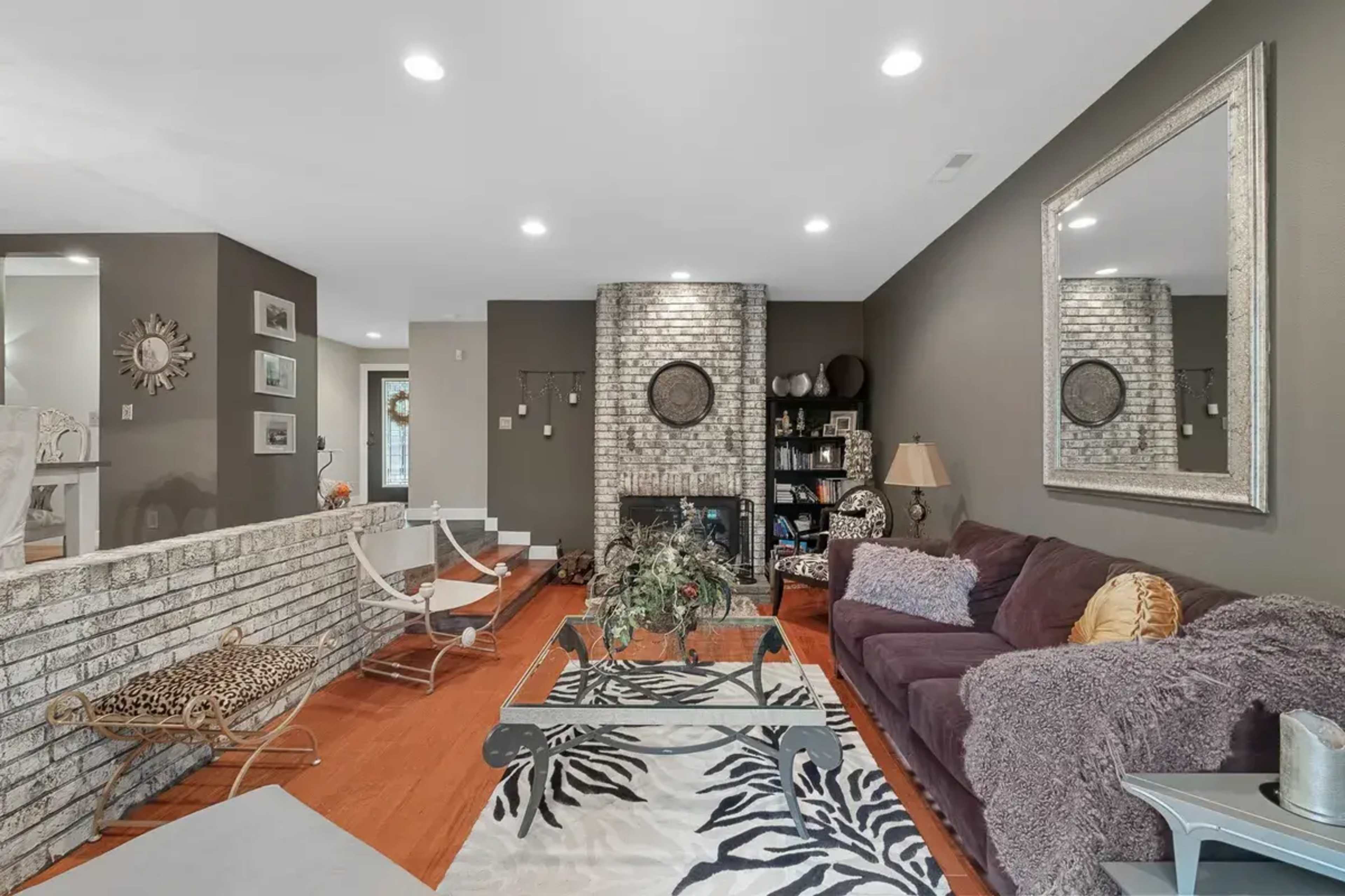 A living room with a brick fireplace, a dark sofa, and a patterned area rug, featuring shelves and decorative items in the background.