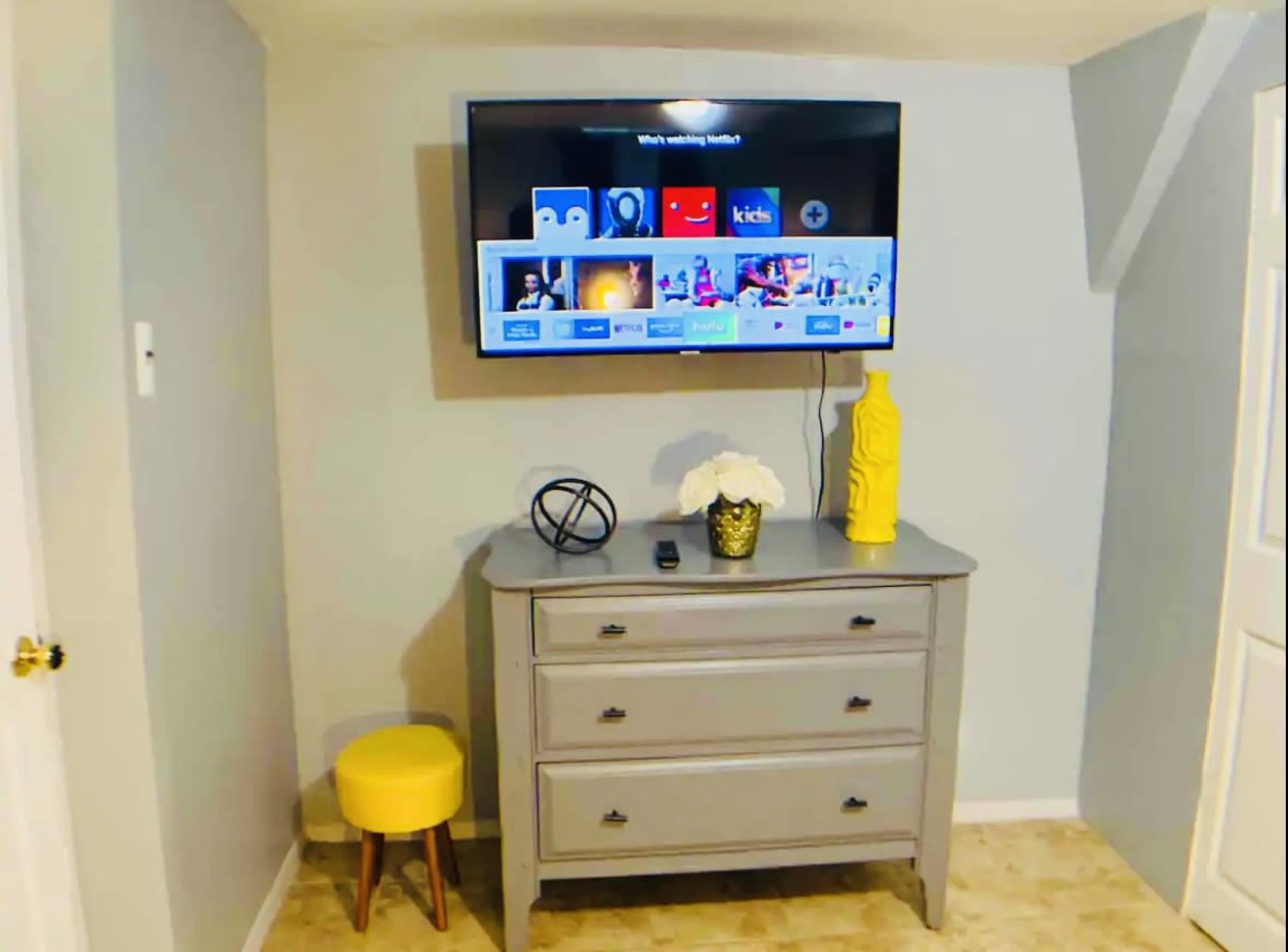 A wall-mounted TV displays various menu options above a gray dresser with a yellow decorative vase and a small yellow stool nearby.