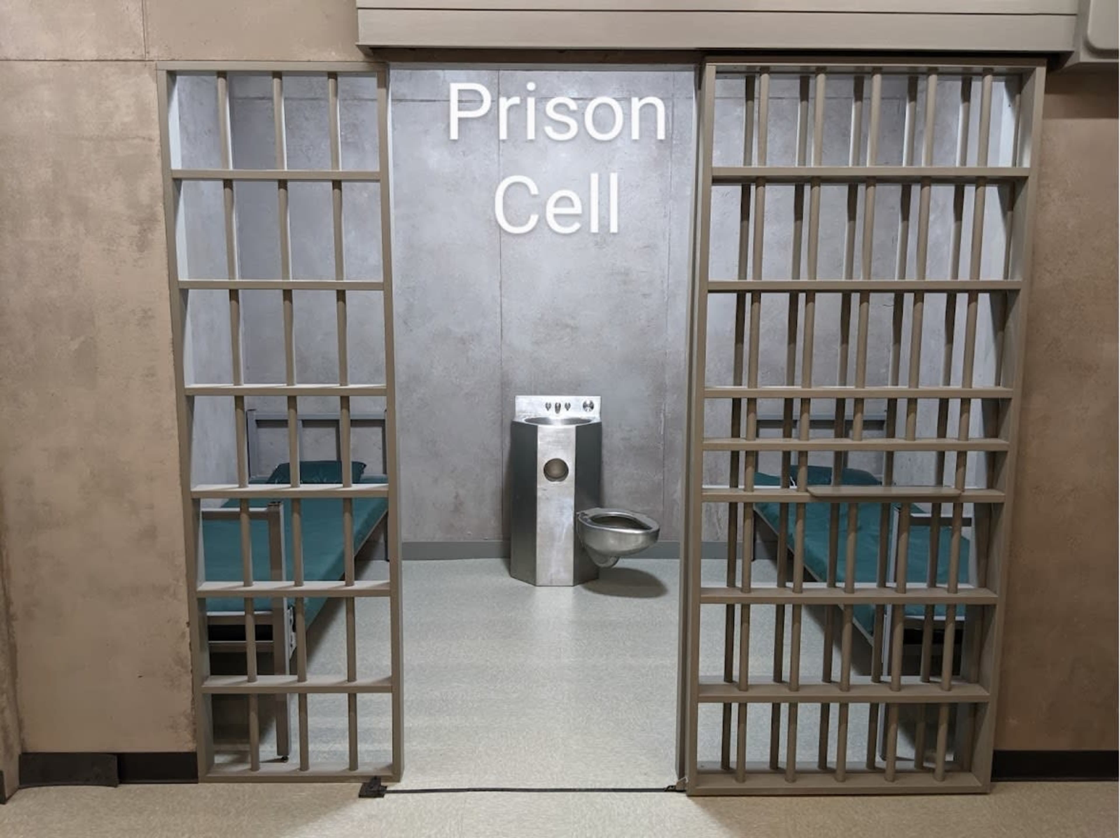 The image shows a prison cell with two beds, a stainless steel toilet, and metal bars at the entrance.