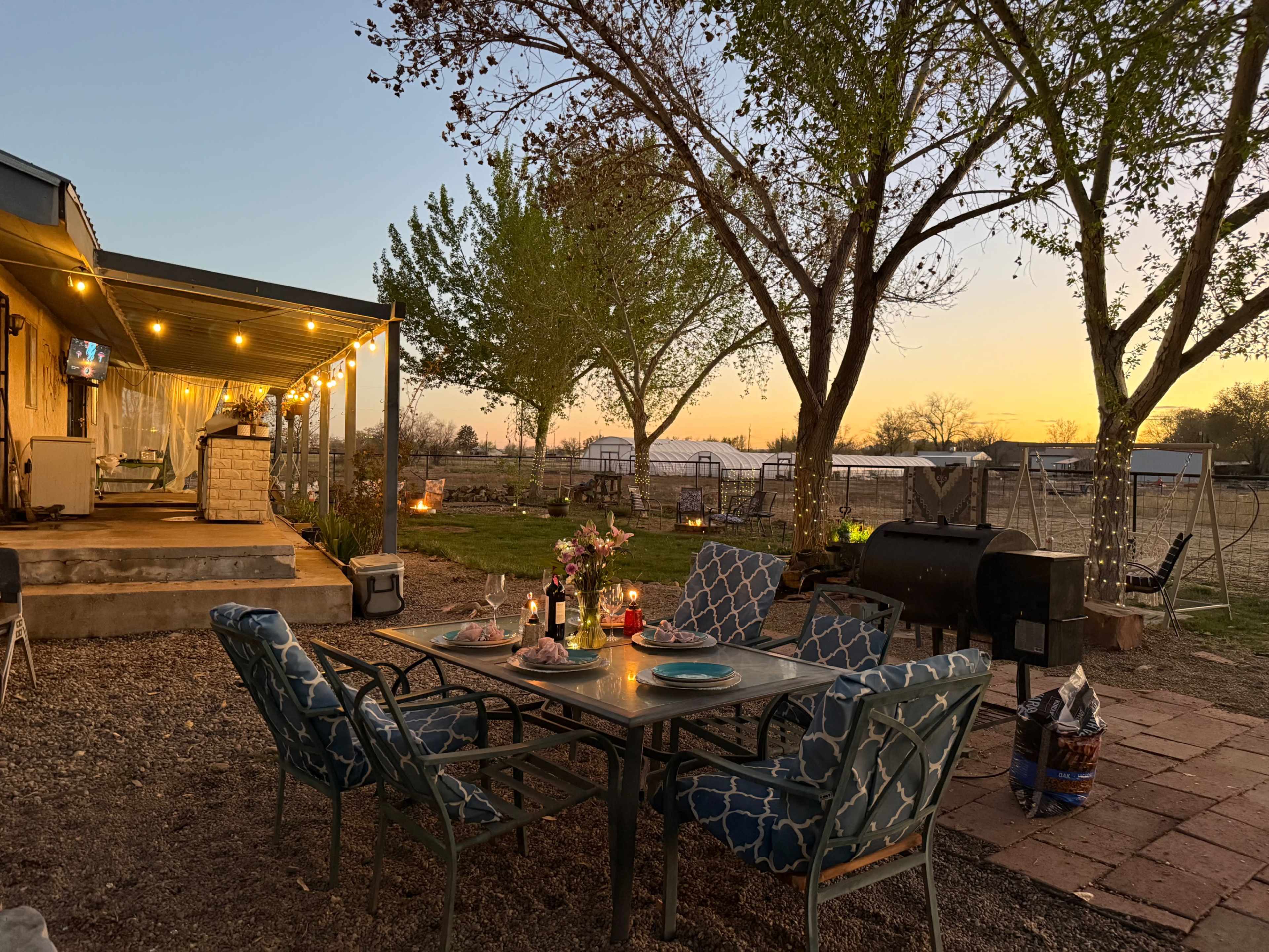 The image shows an outdoor dining area with a table and chairs under string lights, surrounded by trees, at sunset.