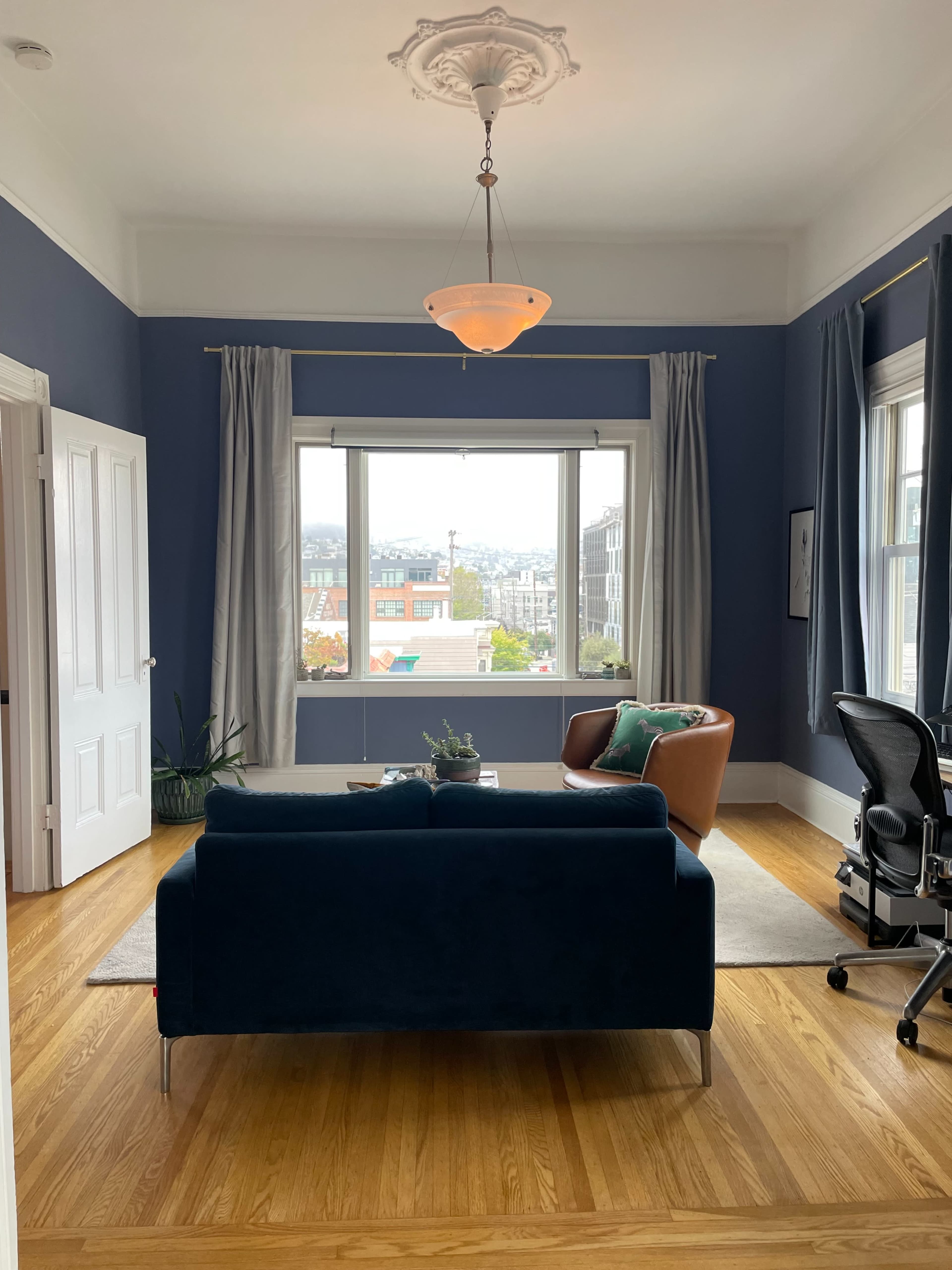 The image shows a styled living room featuring a blue couch, a brown armchair, and a view of the city through large windows.