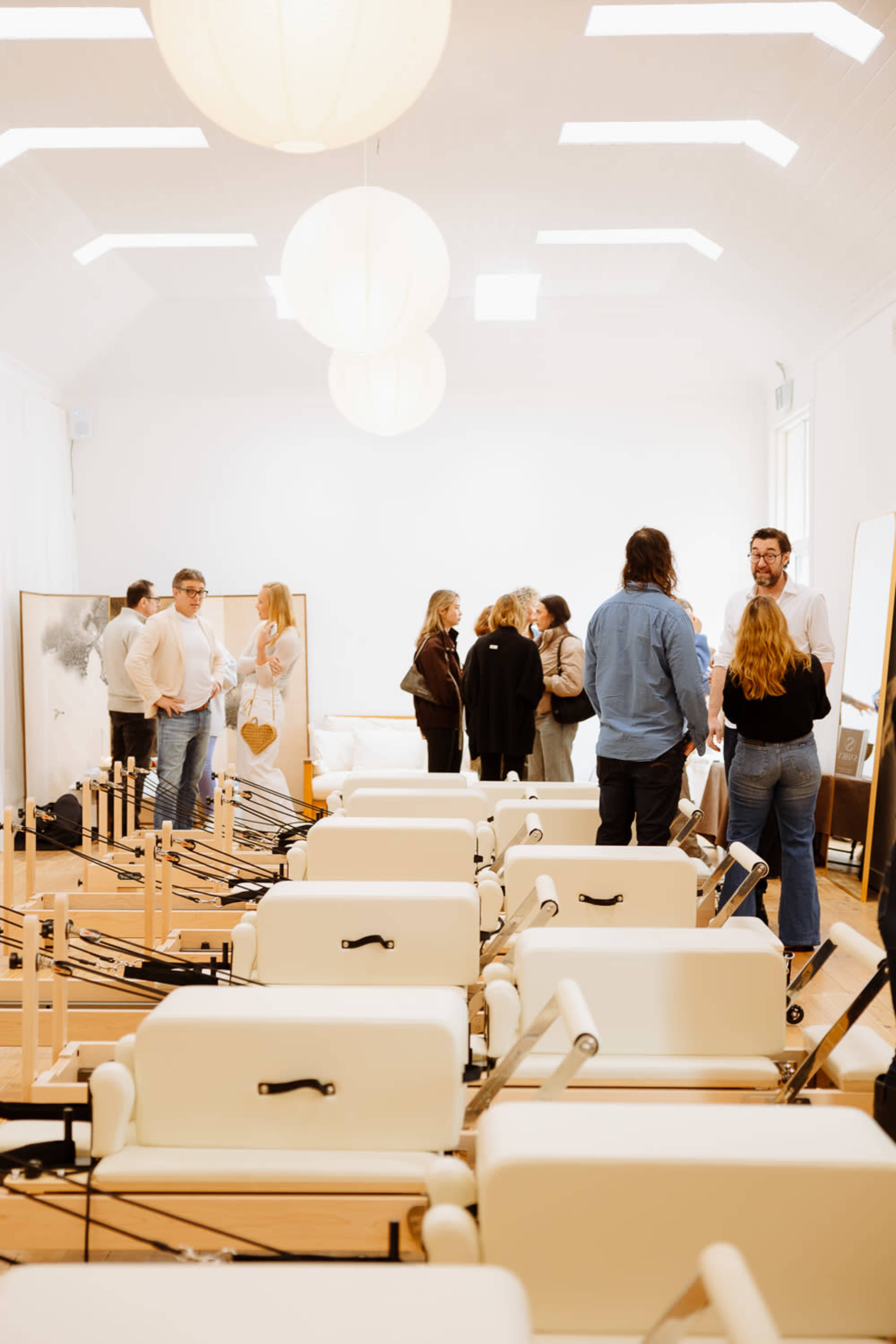 A group of people talks and mingles in a bright room filled with reformer Pilates equipment.