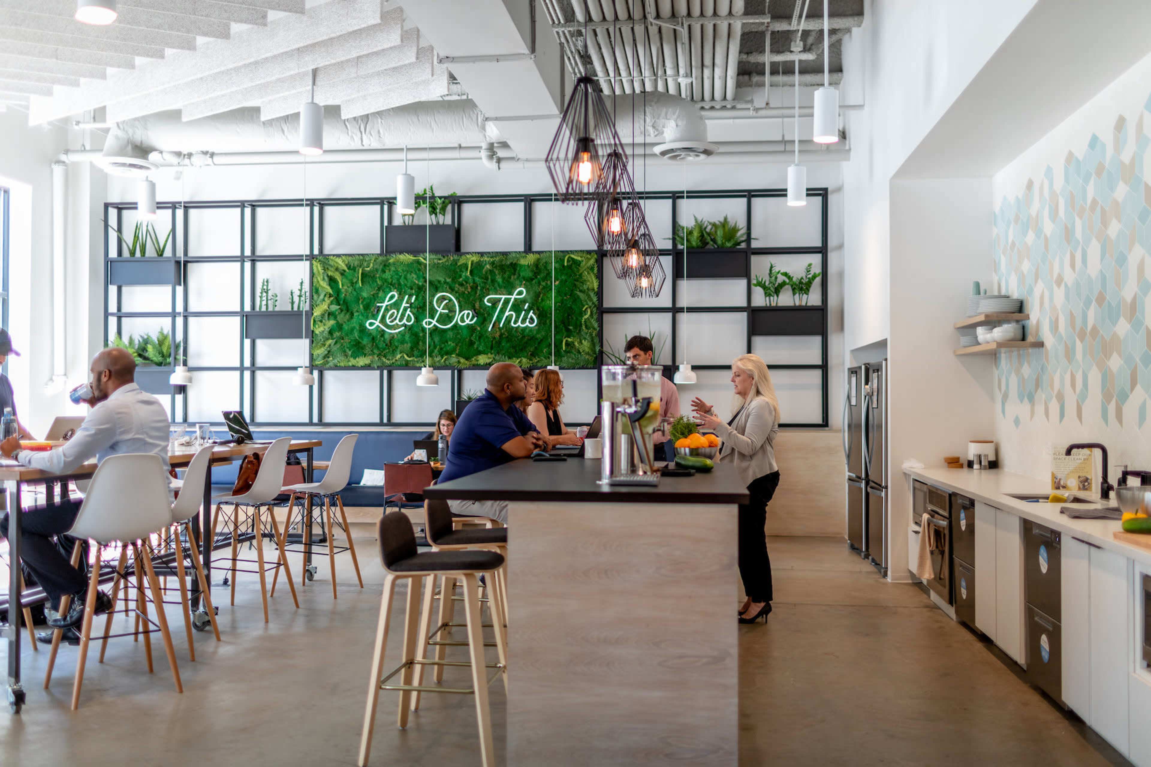 The image shows a modern office space with a bar area and seating arrangements, featuring greenery on the walls and people engaging in conversation.