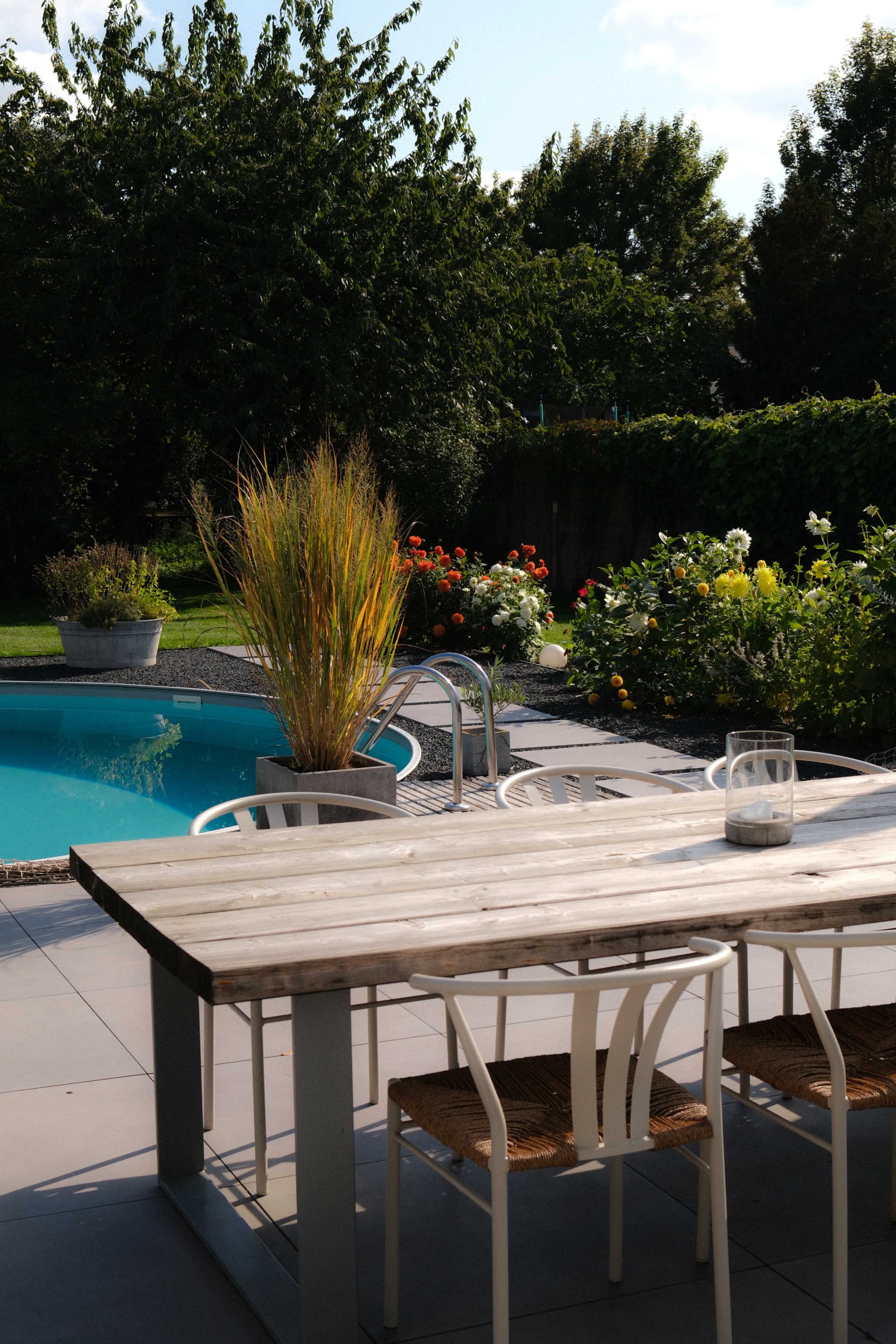The image shows a patio area with a wooden dining table and chairs, overlooking a swimming pool surrounded by vibrant greenery and flowers.