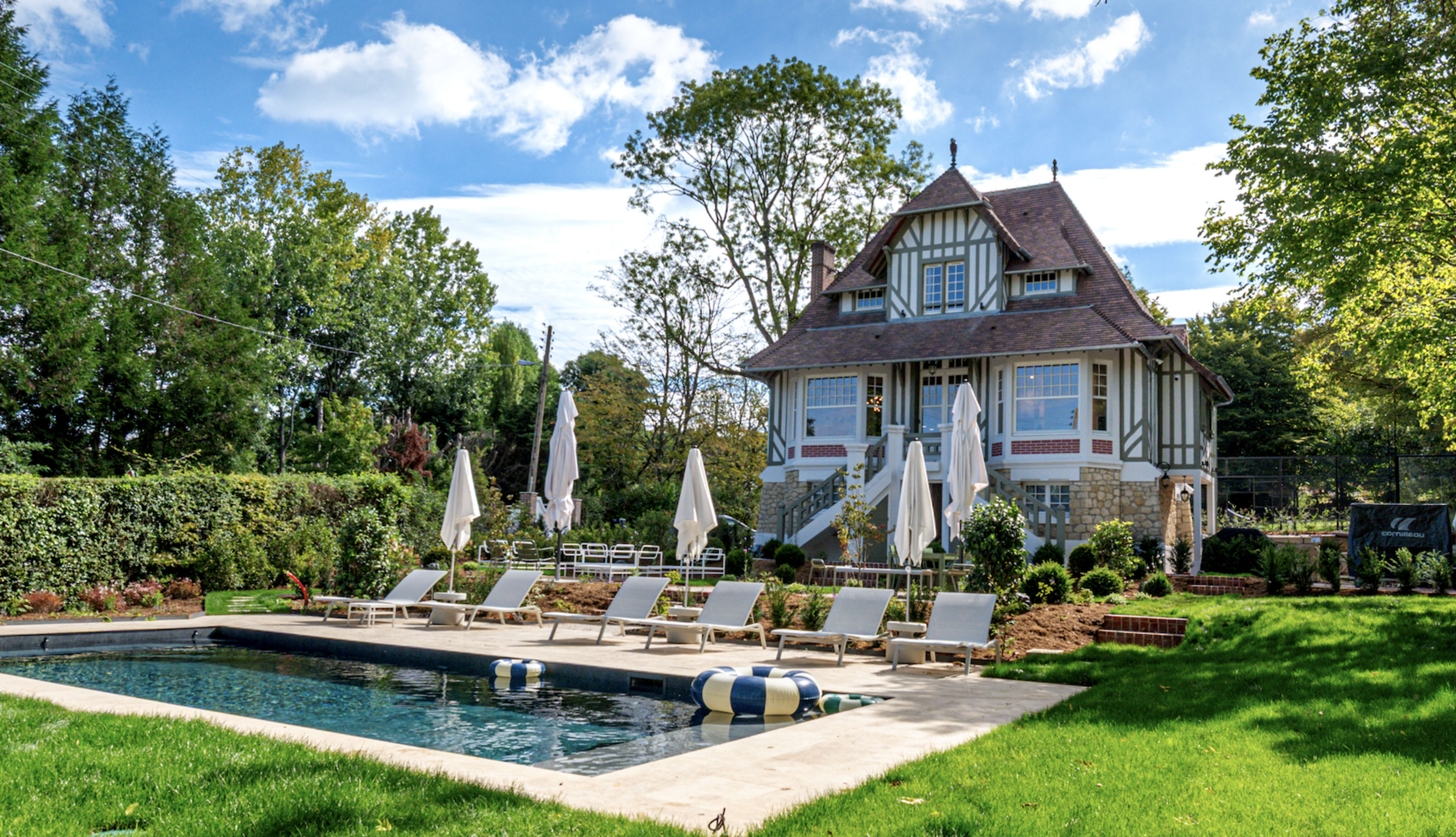 A large, two-story house with a steep roof and gabled dormers sits beside a swimming pool, surrounded by lounge chairs and landscaped greenery.