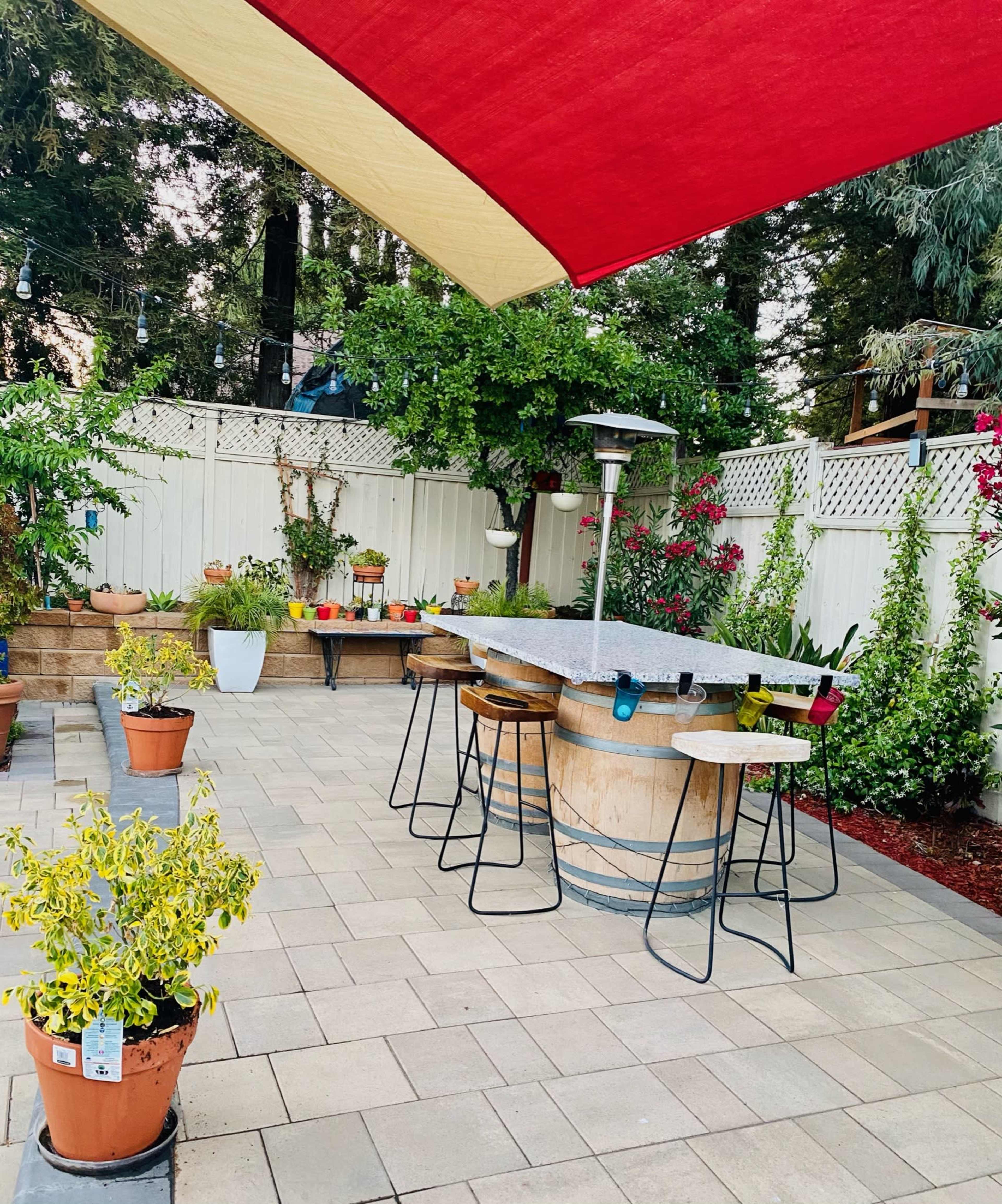 A patio area features a circular stone table with bar stools, surrounded by potted plants and a shaded awning.