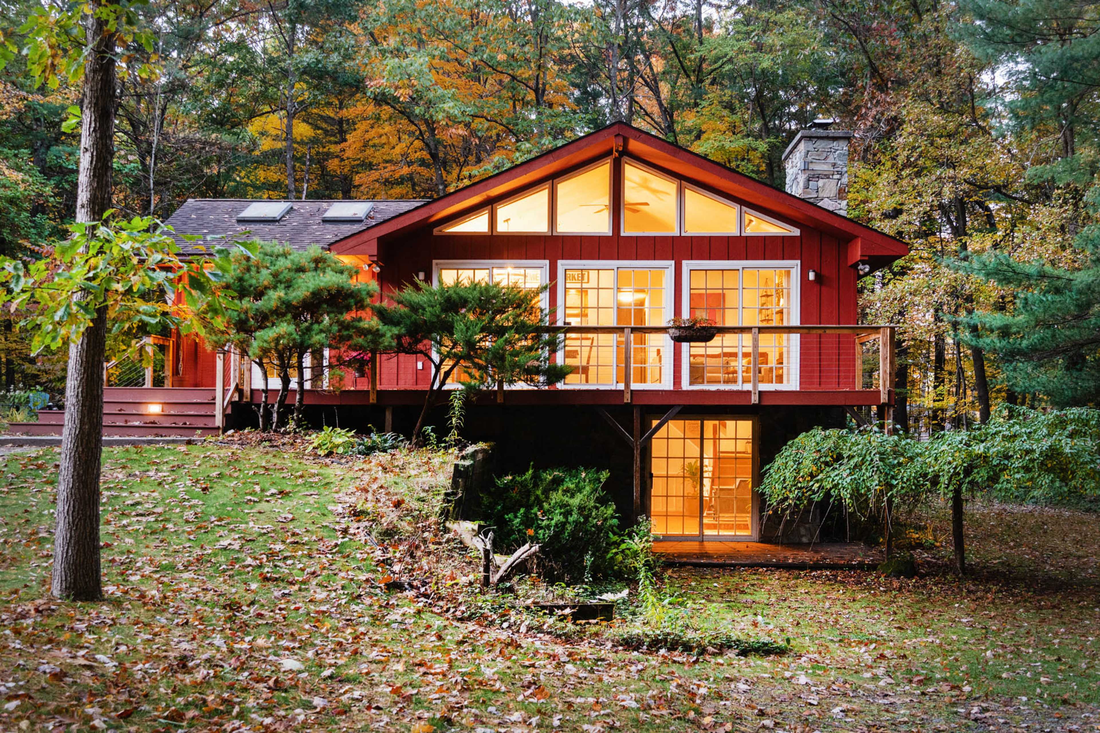 A red cabin with large windows overlooks a landscaped yard surrounded by trees displaying autumn foliage.