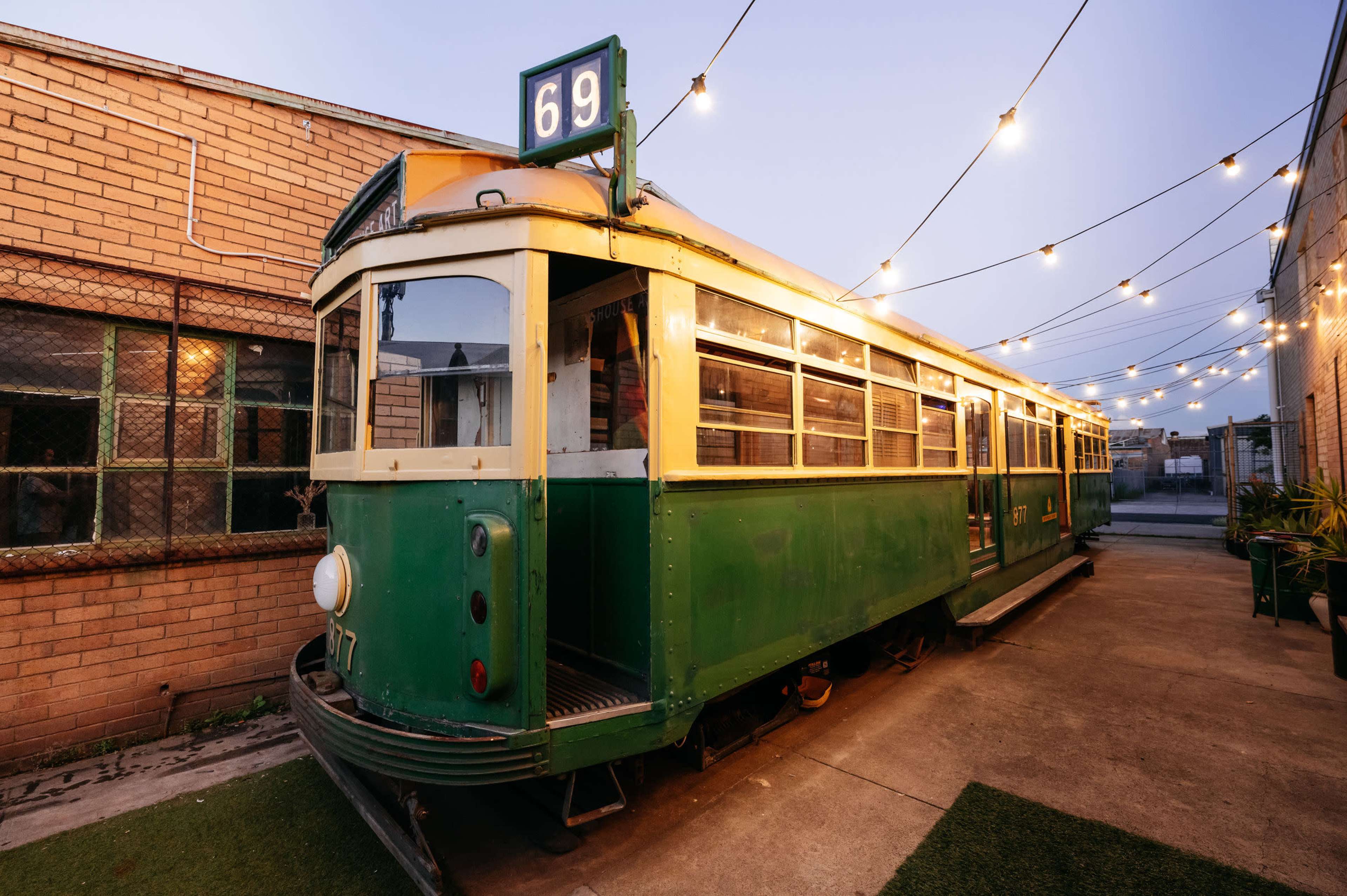 A vintage green tram is parked along a pathway illuminated by string lights at dusk.