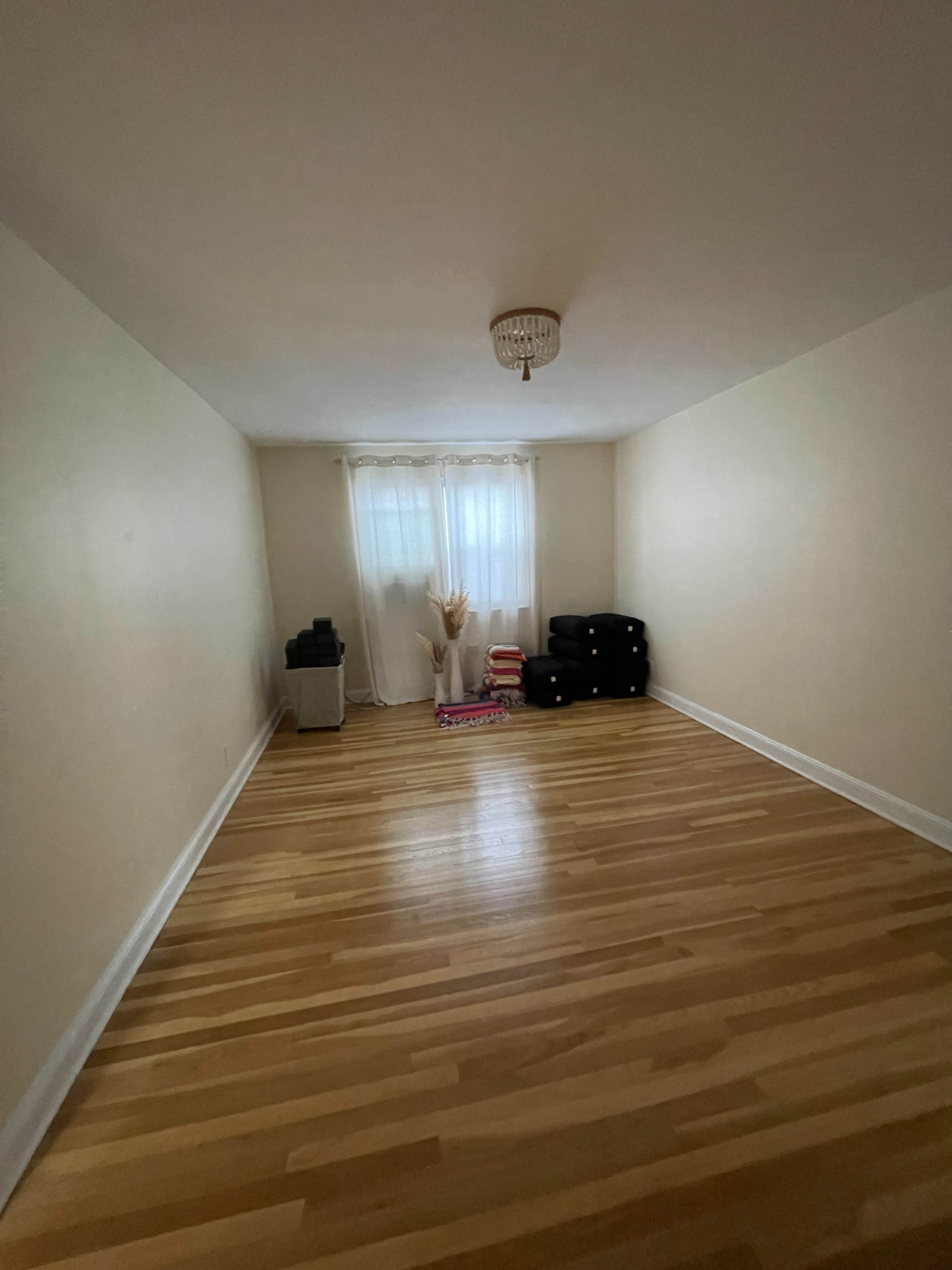 An empty room with wooden flooring, white walls, and a window covered by sheer curtains, accented by a small decorative arrangement and some stacked items in one corner.
